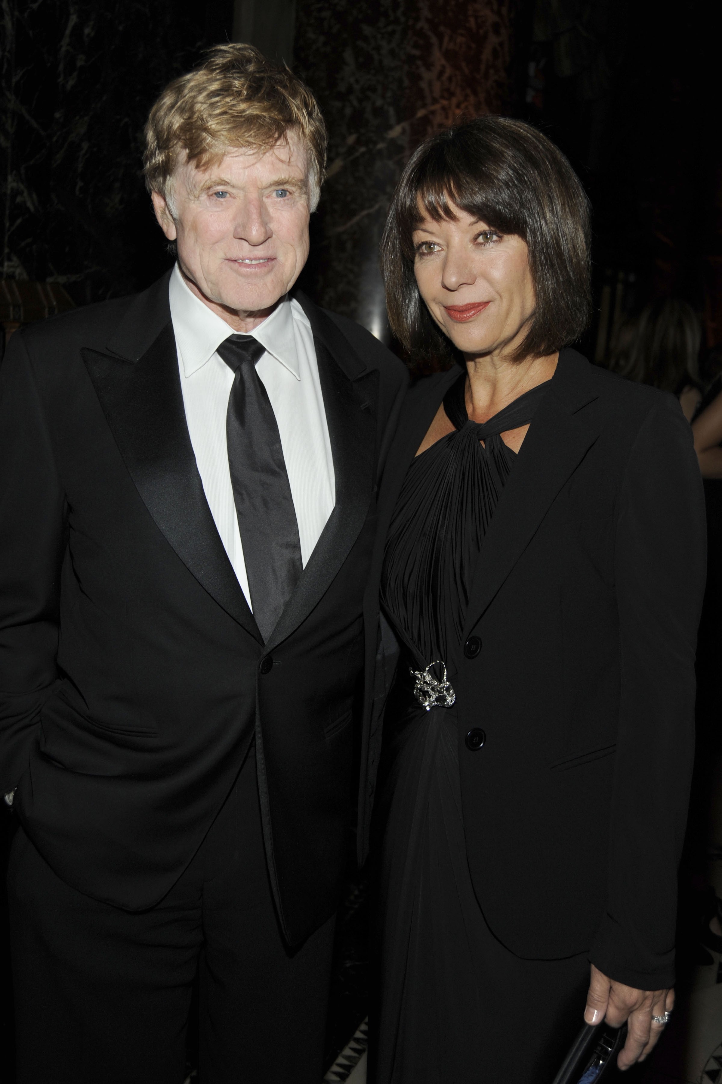 Robert Redford and Sibylle Szaggars-Redford appear at the Americans for the Arts National Arts Awards in New York City on October 5. Szaggars wears a black gown with a halter neckline, a matching tailored jacket, and a jeweled brooch detail at the waist. Redford wears a classic black tuxedo with a white shirt and black tie. They pose together in front of a step-and-repeat backdrop.
