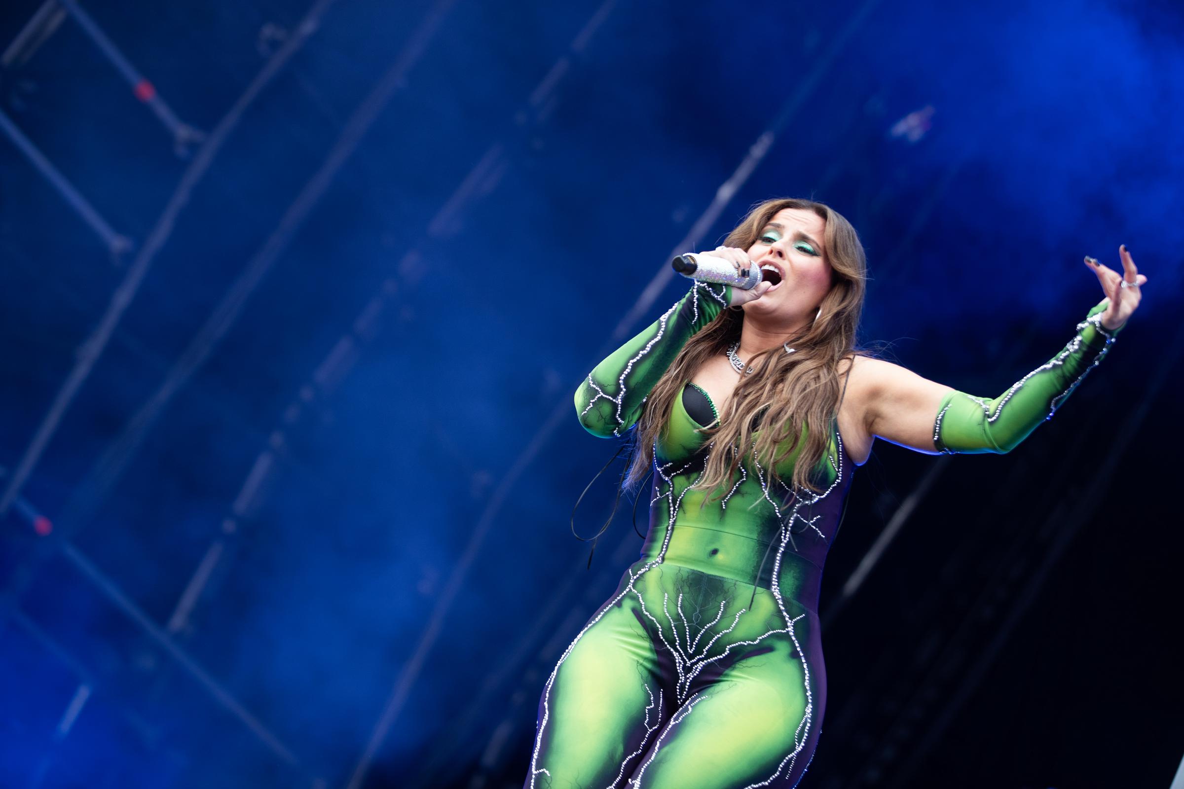 Furtado performs in a green and black bodysuit with silver embellishments and cutout detailing. She holds a microphone in one hand while singing, with blue stage lighting behind her.