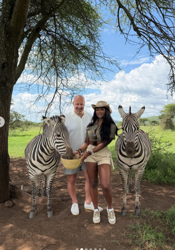 Framed by towering trees, Joe McCann and Ashly Robinson stand between two zebras, casually feeding them. | Source: Instagram/ashleejenae