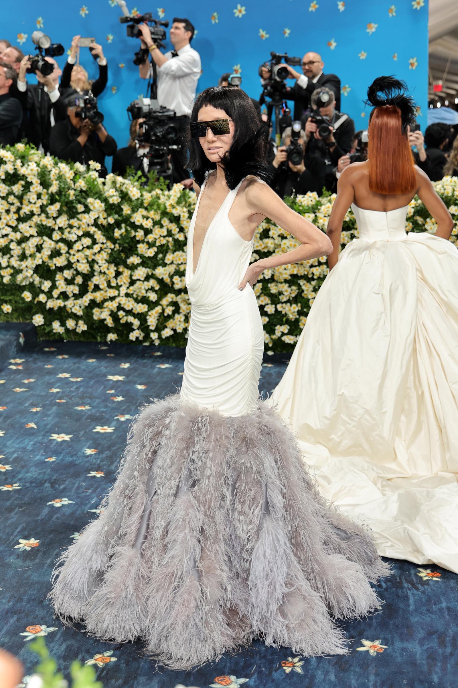 With cameras flashing behind her, Vera Wang poses in profile, showcasing the gown's sculpted draping and voluminous feathered skirt against a floral backdrop.