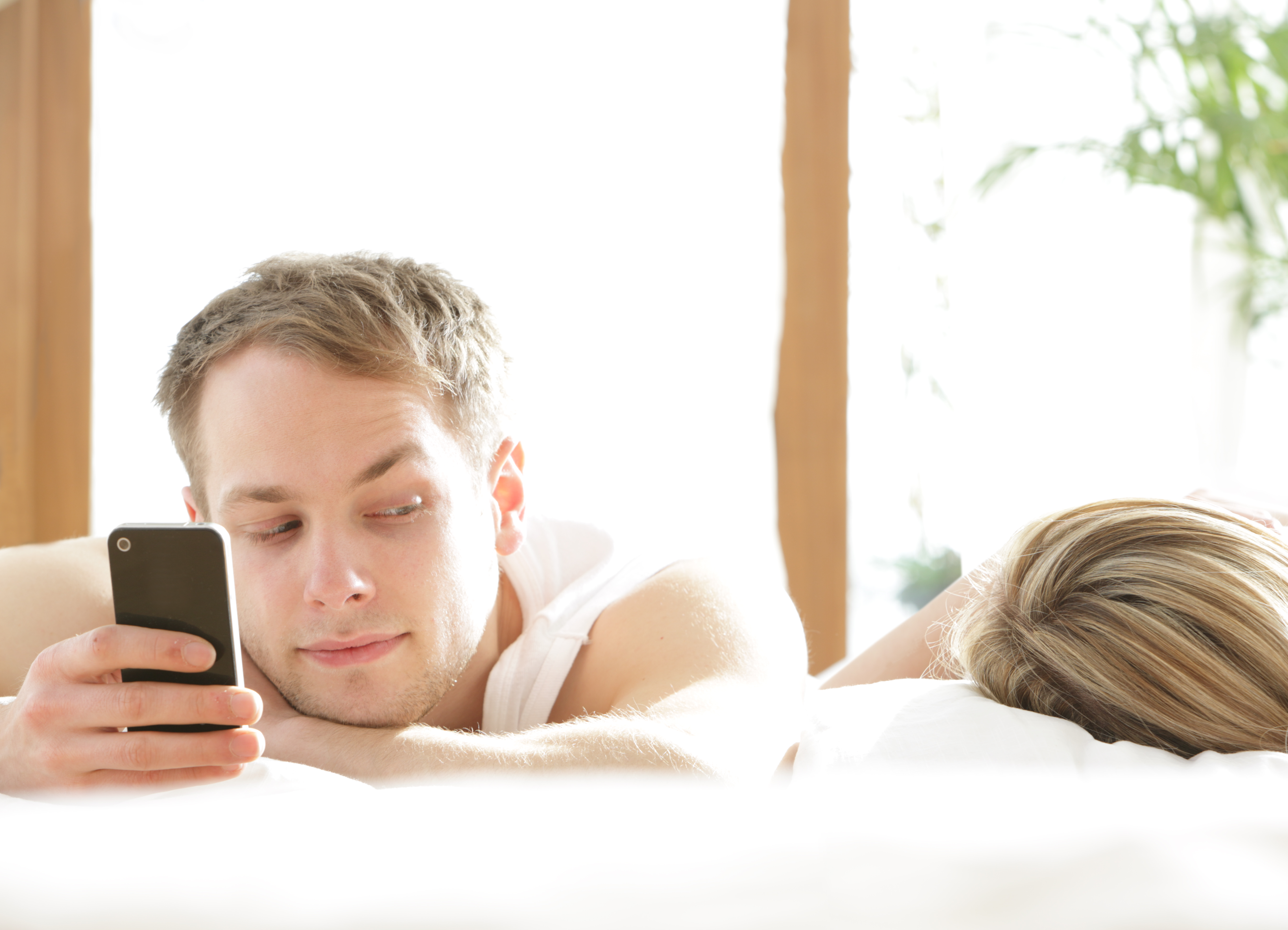 Man with phone in bed, looking at woman asleep | Source: Getty Images