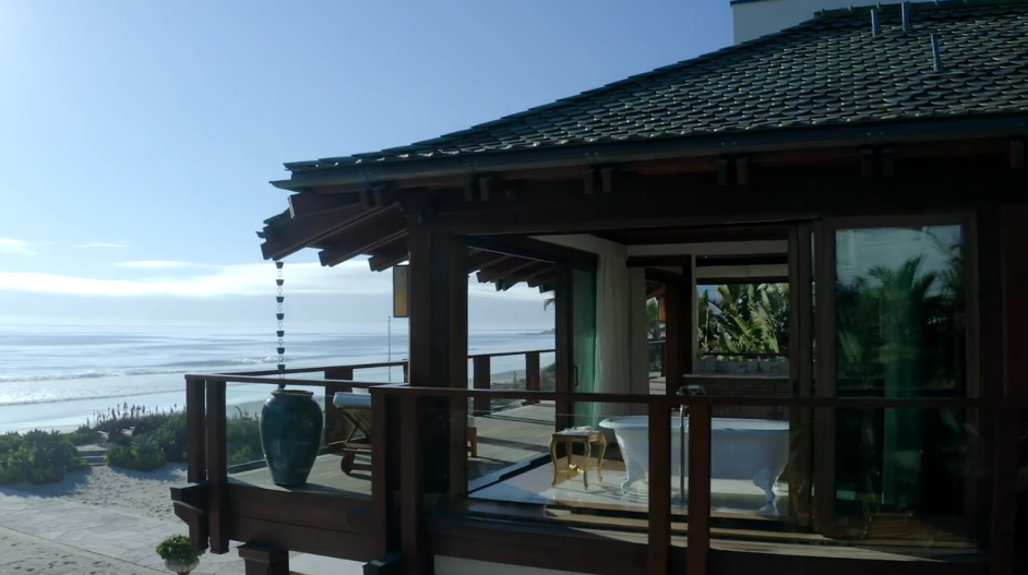 Perched on the edge of the second-story balcony, the primary bathroom suite commands sweeping views of the Malibu ocean and sandy beach. The combination of open timber framing, a green clay-tiled roof, and retractable glass walls enhances the home's seamless blend of indoor and outdoor resort living. | Source: YouTube/Architectural Digest