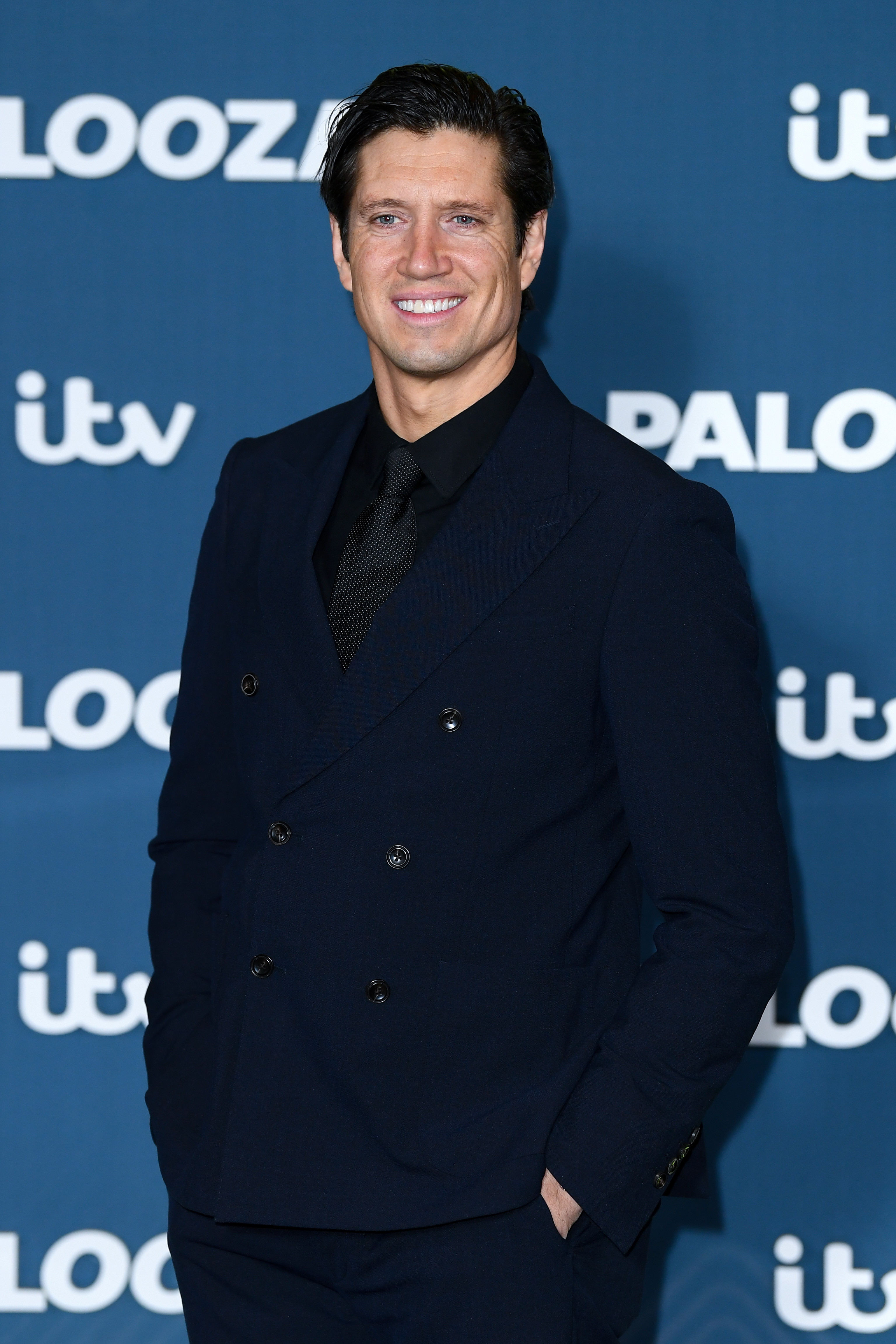 Vernon Kay cuts a sleek figure in a dark double-breasted suit with a black shirt and tie, smiling softly as he poses against a blue backdrop.