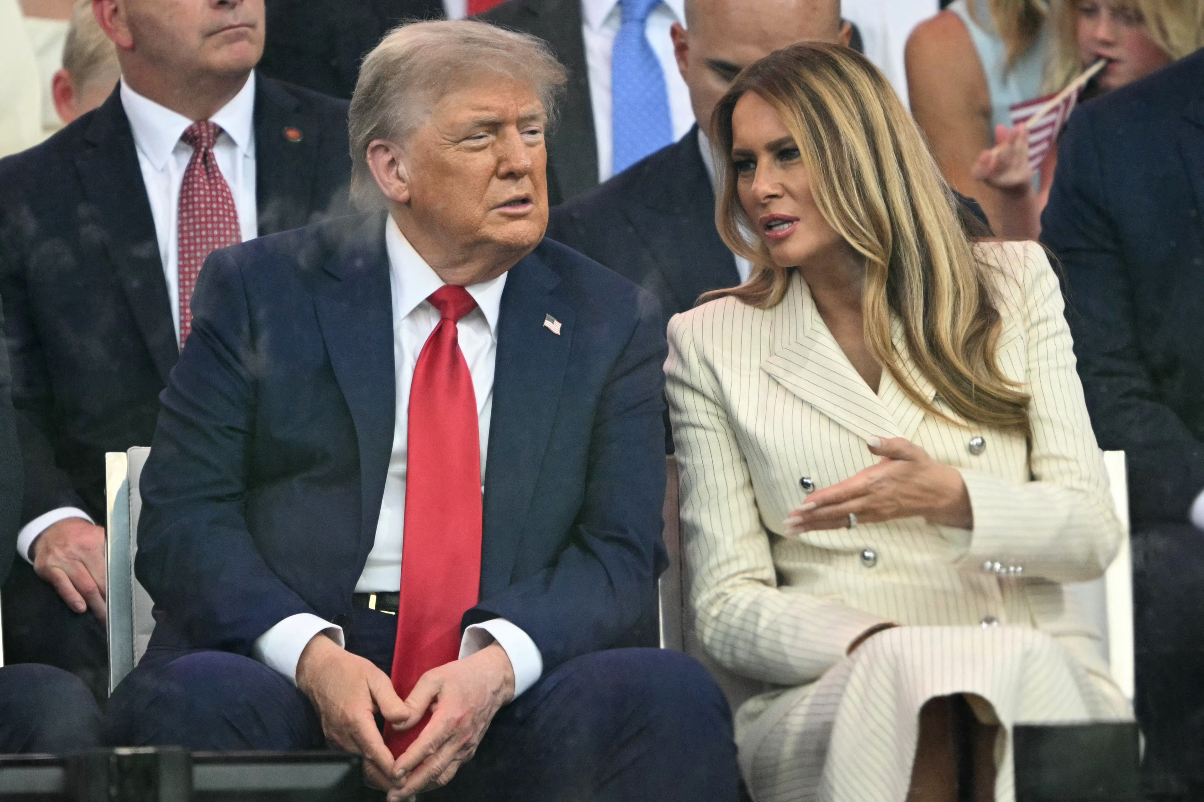 Seated closely, Donald and Melania Trump appear to be mid-conversation. Melania gestures with one hand as she speaks, while Trump listens intently. Their exchange takes place during the military parade in a rare candid moment.