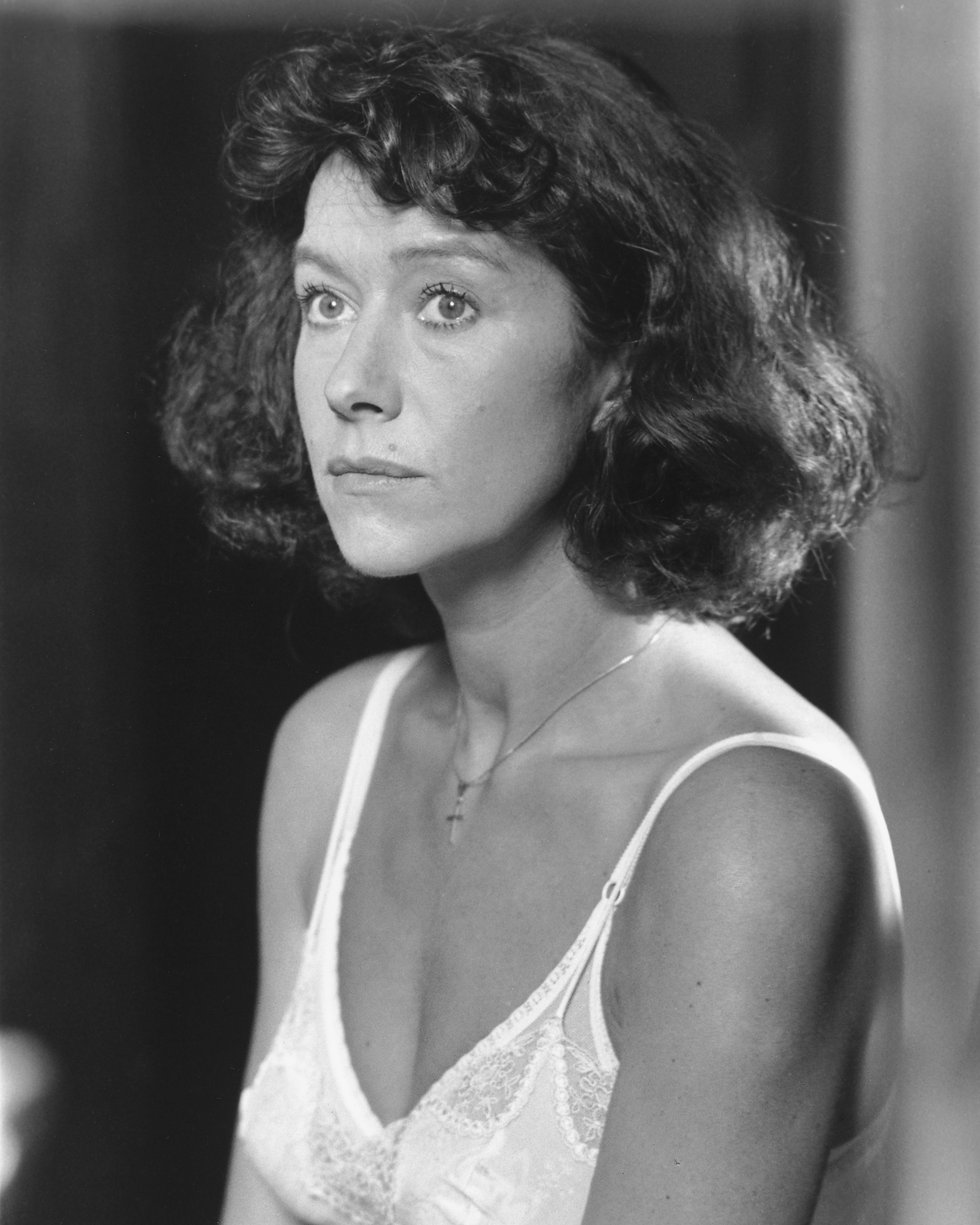 Helen Mirren appears in a contemplative moment, captured in a soft-lit black-and-white portrait, wearing a delicate lace camisole and a simple cross necklace, her expression serene and her curled hair framing her face with quiet intensity.