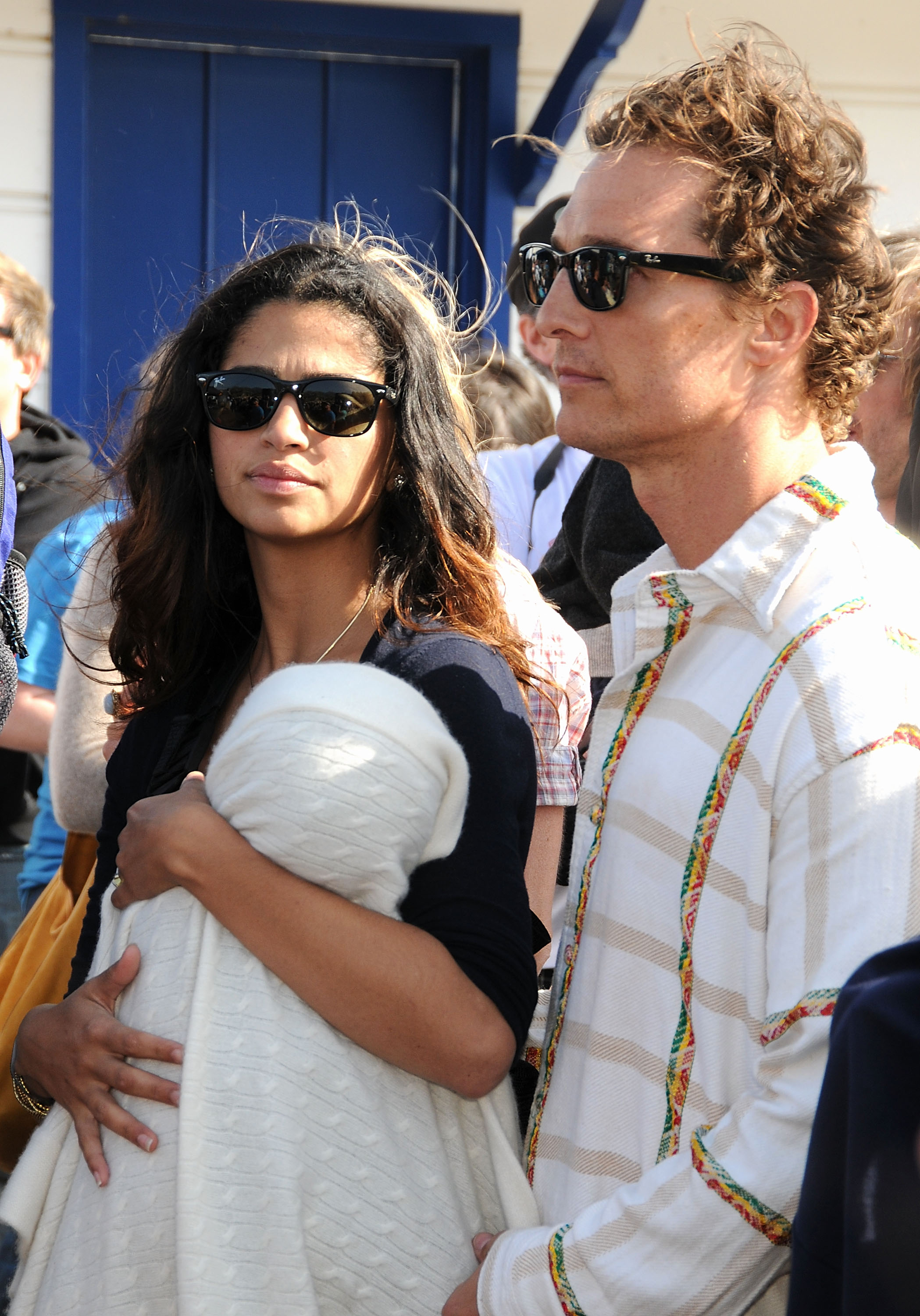 Levi Alves McConaughey is held close against Camila Alves, wrapped in a white blanket. Camila Alves wears a dark top and sunglasses, while Matthew McConaughey stands beside her in a patterned shirt and sunglasses. They stand together in a crowded outdoor setting.