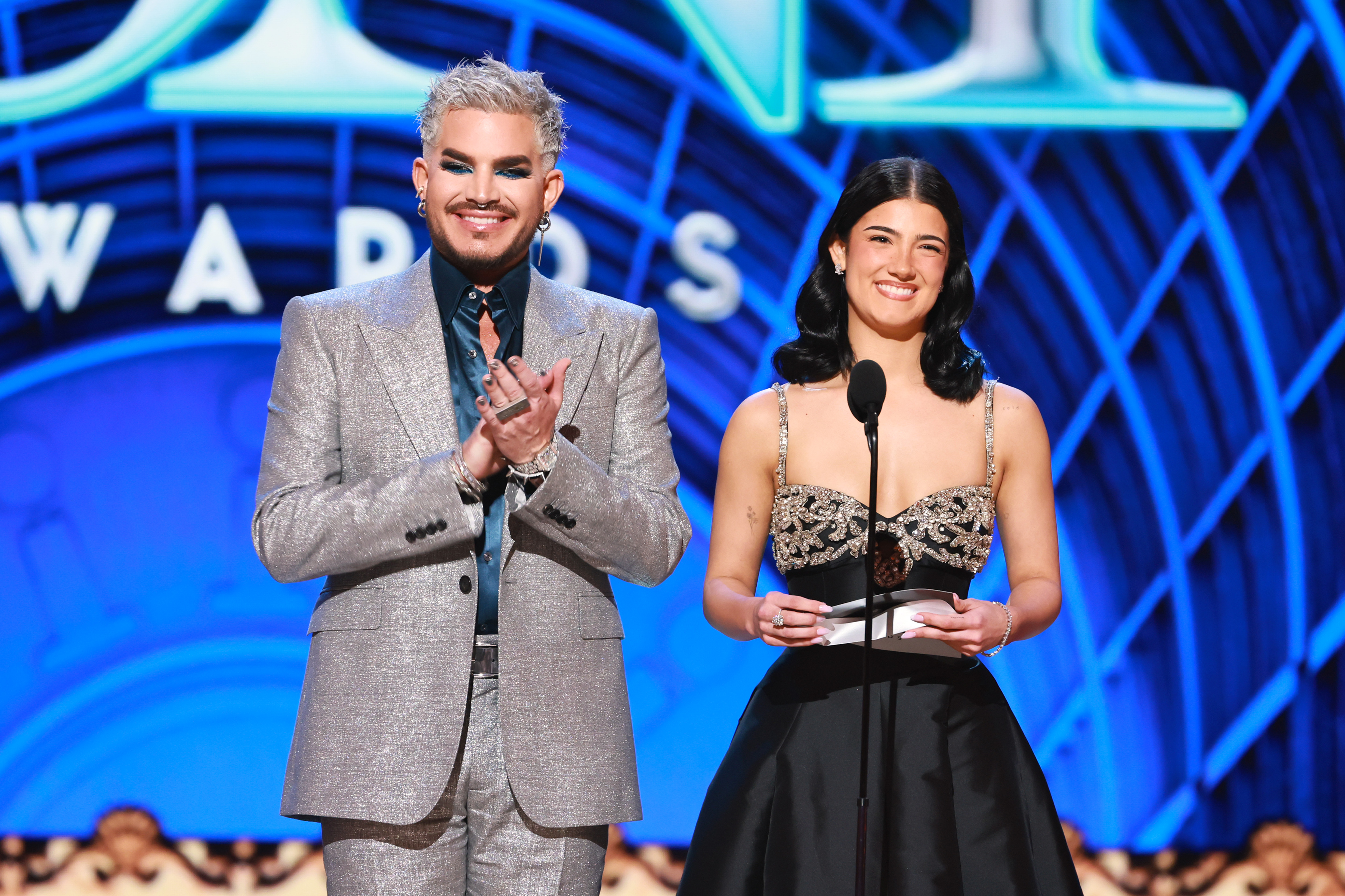 Adam Lambert and Charli D'Amelio exchange a glance and a smile as they stand at the mic. His glittering silver suit and bold makeup pair strikingly with her sleek, strapless gown, creating a polished moment of style and stage chemistry.
