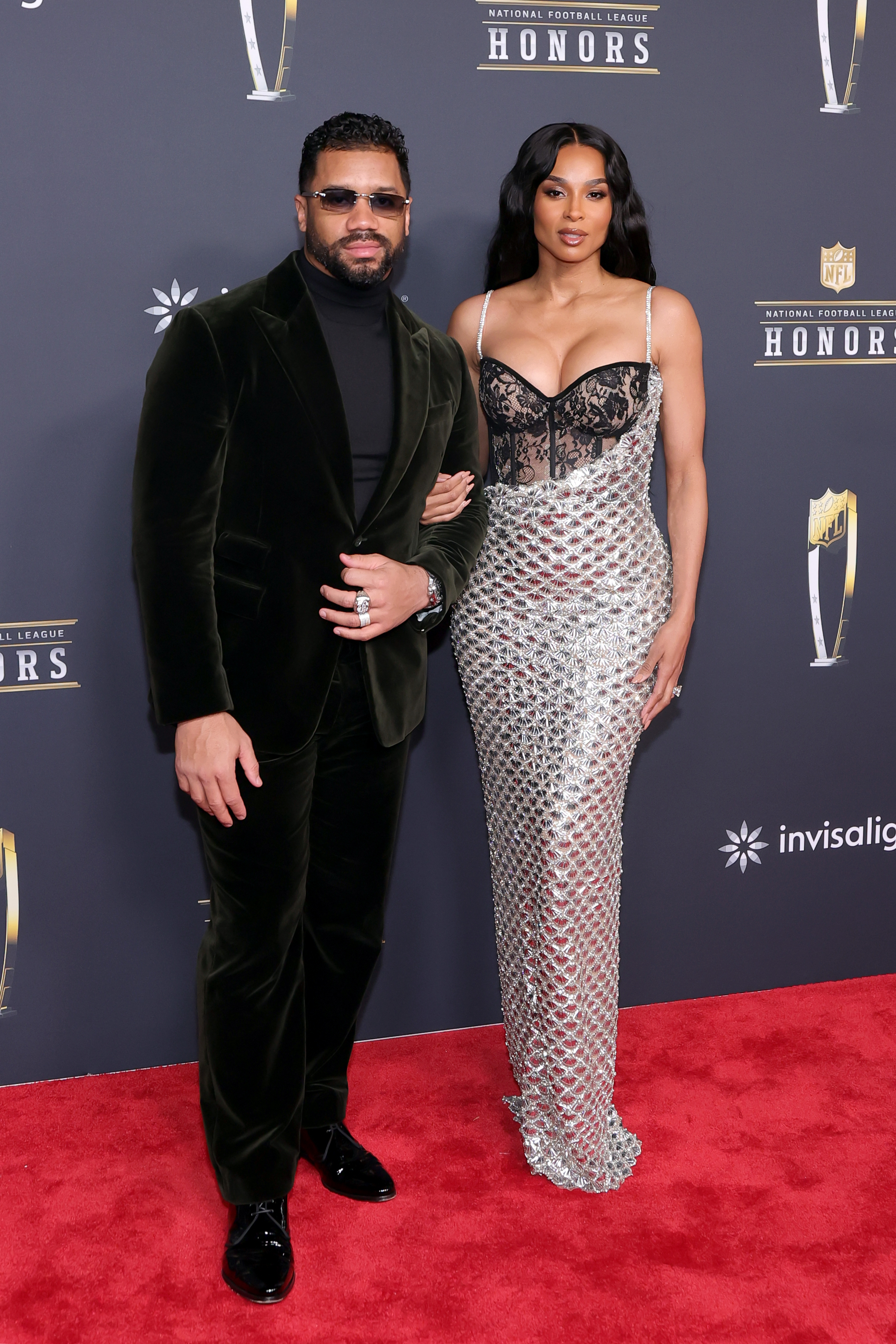 Russell Wilson and Ciara walk the red carpet arm in arm, Russell composed in a tailored look as Ciara smiles in a form-fitting metallic dress, blending elegance and star power in a polished appearance.