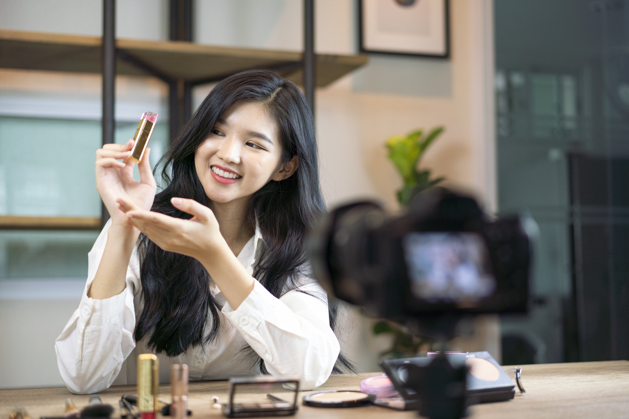 A young Asian female sits in front of personal camera and smiles as she holds up pink lipstick for a vlog video, Thailand | Source: Getty Images