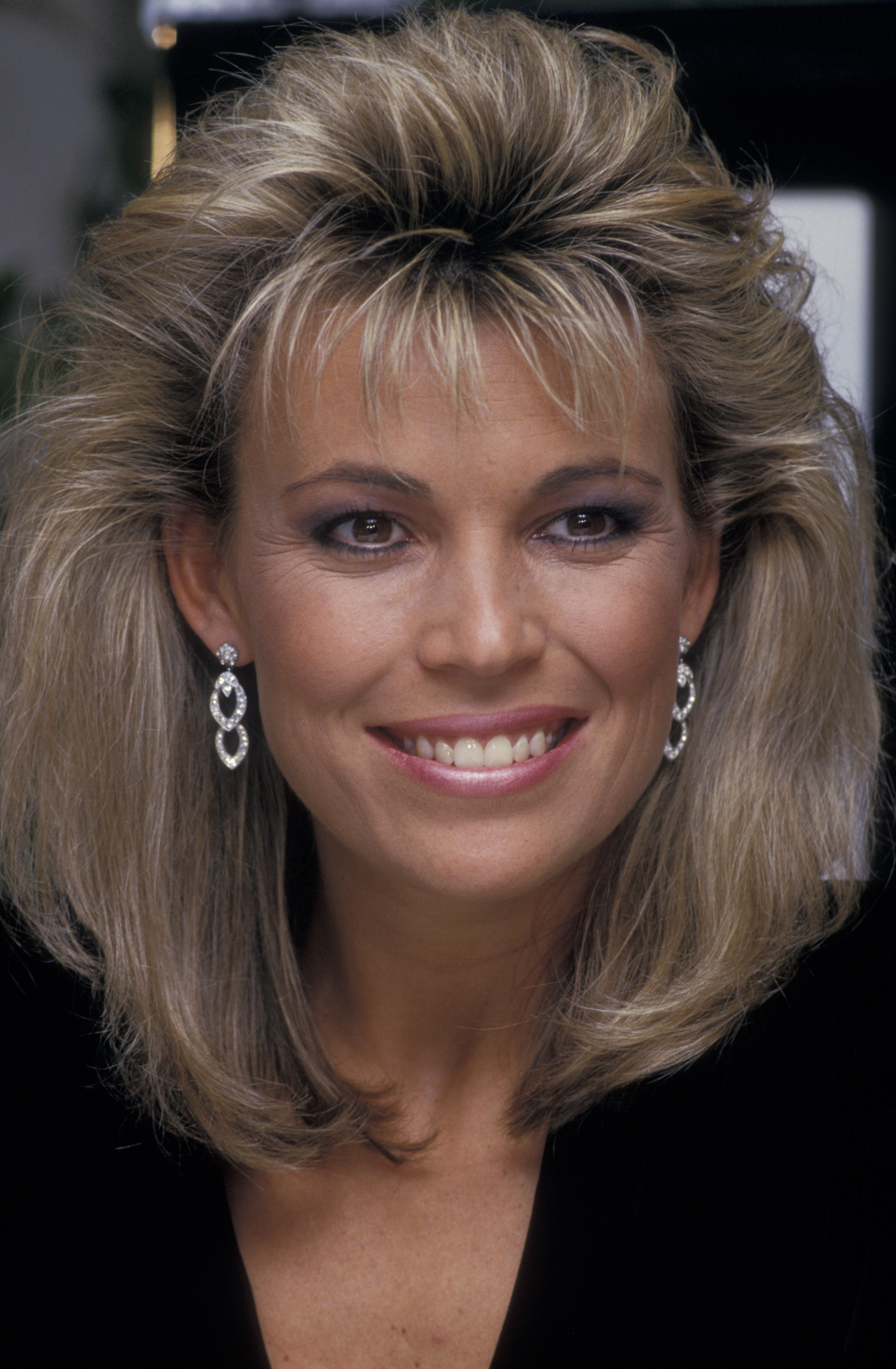 Vanna White smiles in a black outfit with voluminous blonde hair and sparkling earrings while she unveils a clothing line at Bloomingdale's in New York City.