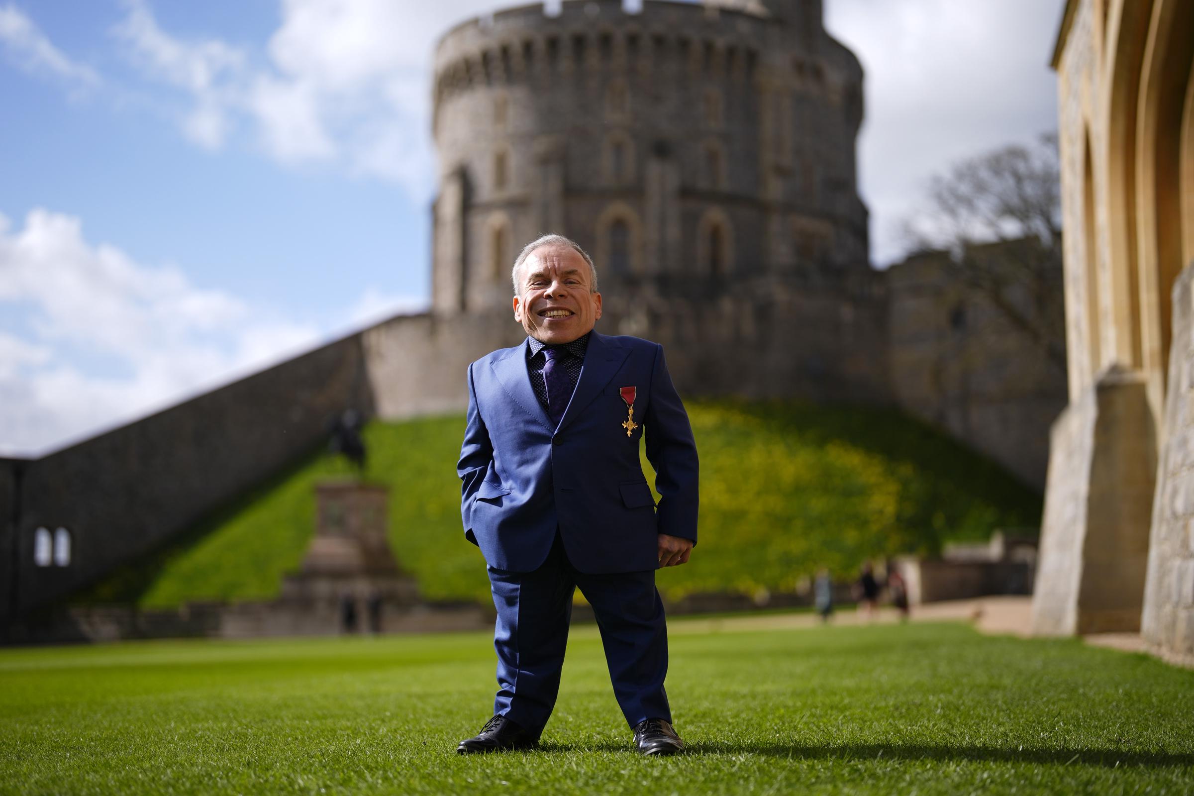 Warwick Davis stands on the castle grounds with a satisfied smile, the ancient stonework of Windsor's Round Tower looming large behind him — a striking image of a man very much at ease with the extraordinary. Named on King Charles III's New Year's Honours list for 2026 alongside Idris Elba and Cynthia Erivo, the 56-year-old actor was recognised on the day for his outstanding services to both drama and charity.