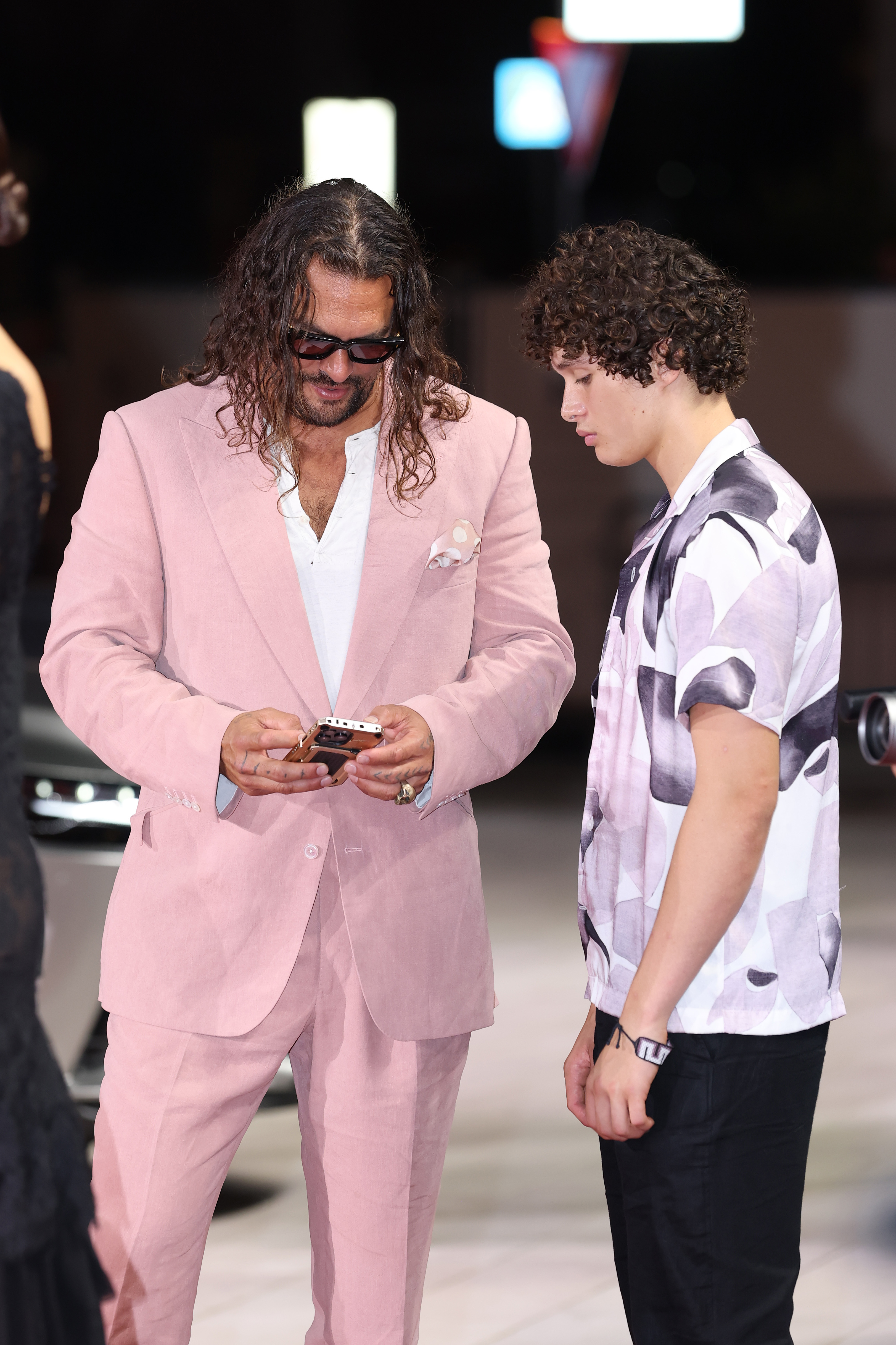 Caught between shots, Jason Momoa glanced at his phone while Nakoa-Wolf Momoa leaned closer, giving the cameras a casual, unscripted moment on the Venice Film Festival red carpet.