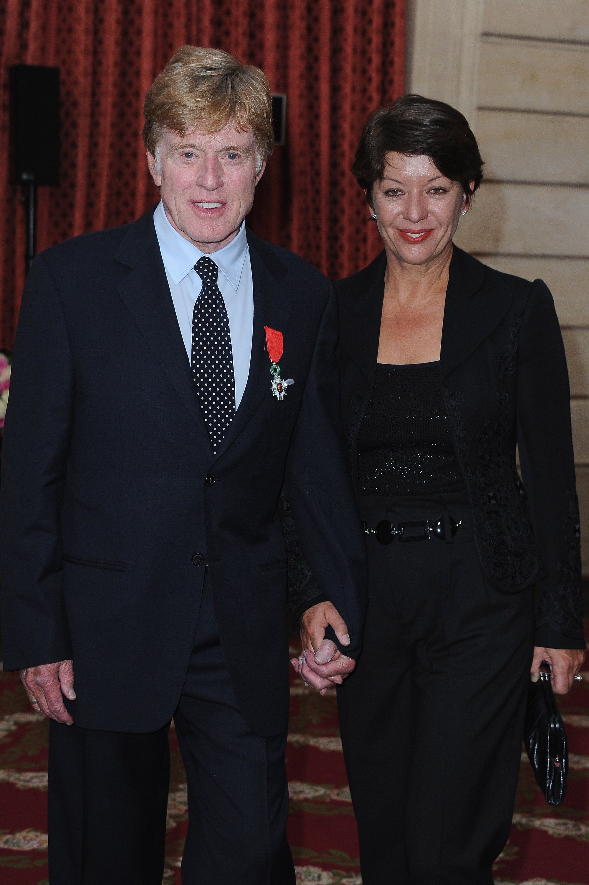The couple is photographed at the Élysée Palace in Paris, France, on October 14, after Redford receives the Legion d'Honneur. Redford wears a dark suit with a white shirt, navy polka dot tie, and the red ribbon and medal on his lapel. Szaggars wears a fitted black jacket with embroidered detailing and a black belt, holding a matching clutch as they stand hand in hand.