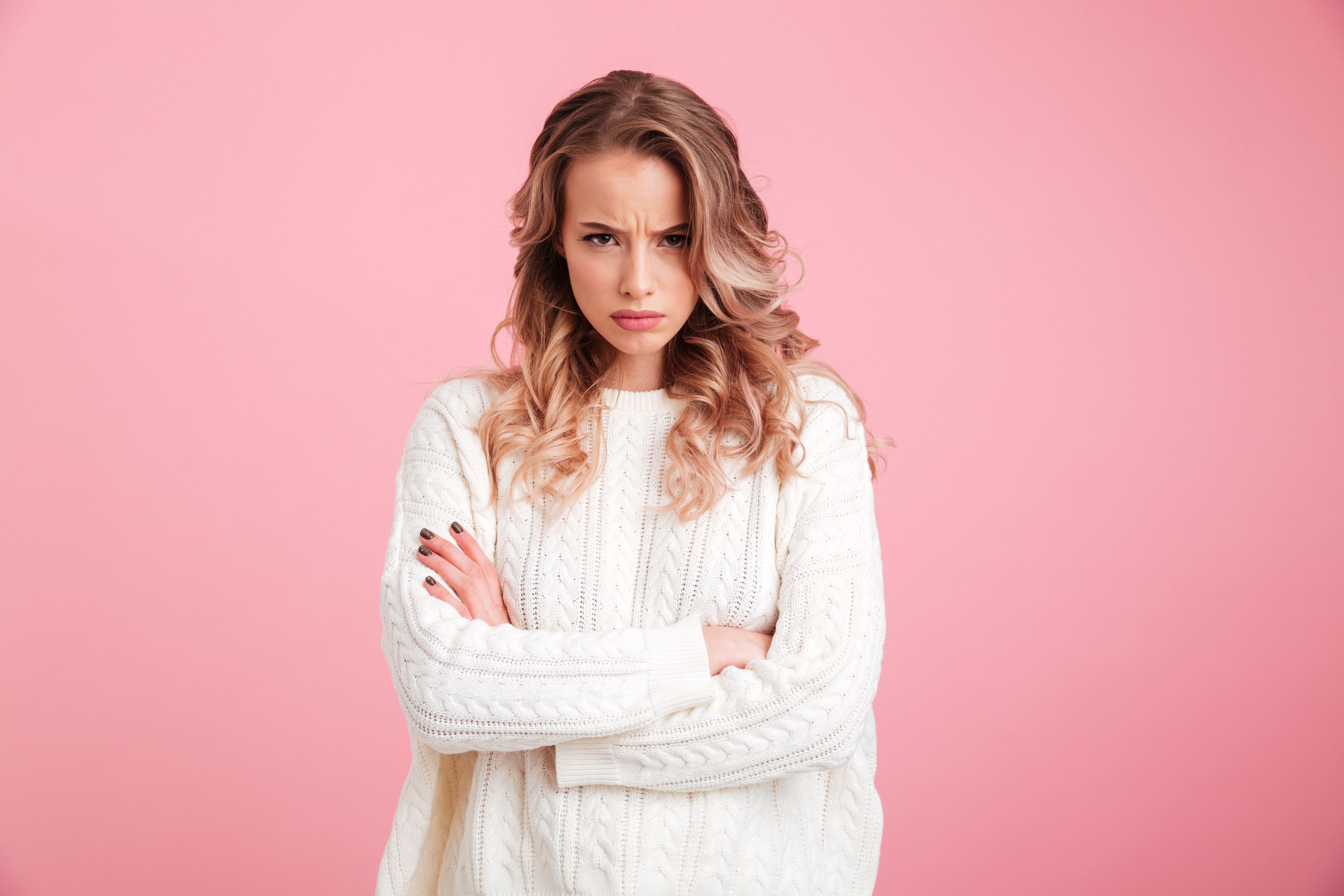 Blonde woman wearing a white jersey in front of a pink background crossing her arms and staring angrily into the camera. | Photo: Shutterstock