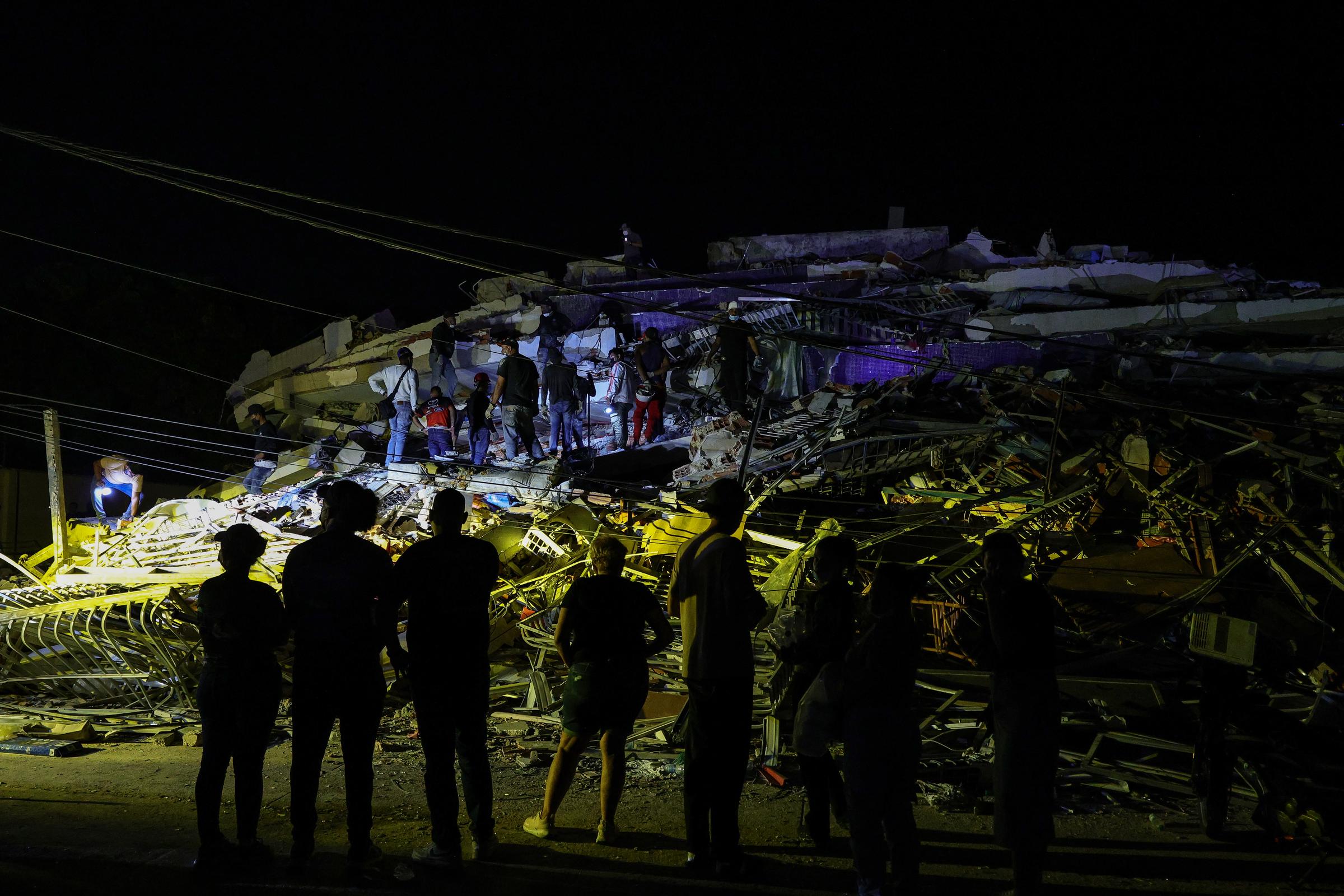 People stand near the collapsed building as rescue crews continue working after dark. Floodlights allowed emergency teams to keep searching through the night. | Source: Getty Images