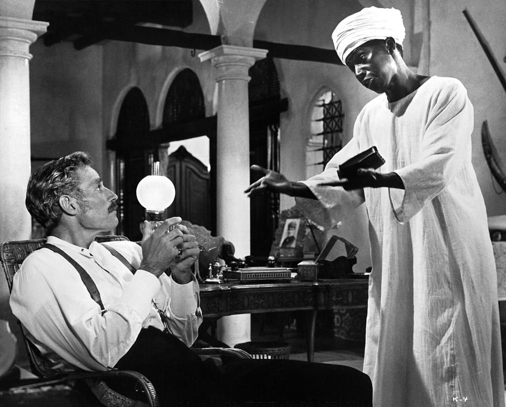 Charlton Heston sitting in a chair looking up at Johnny Sekka who is wearing a turban wrapped around his head in a scene from the film 'Khartoum', 1966. | Photo: Getty Images