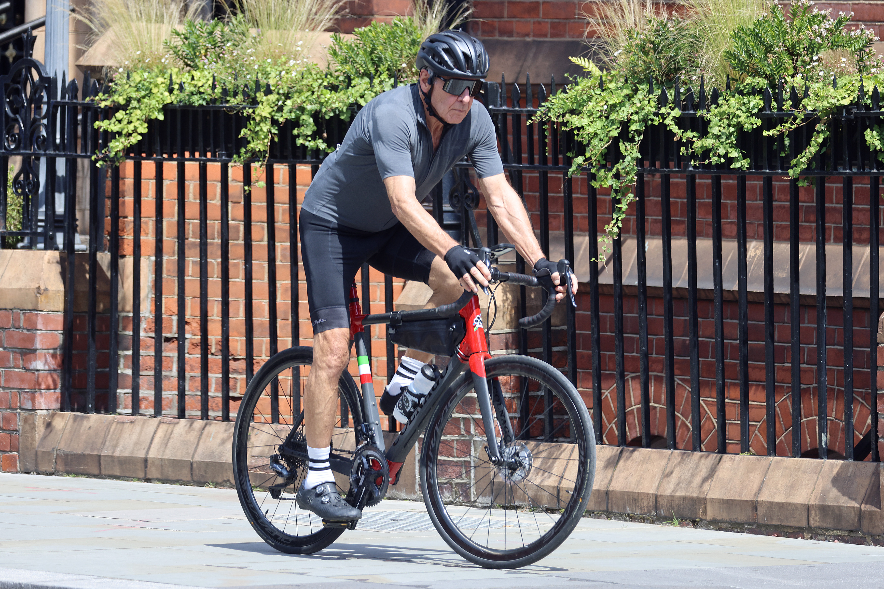 Photographed in July 2021, Ford powers through a street in London's Mayfair district on a red-and-black road bike. Sporting a gray athletic jersey, black cycling shorts, a protective helmet, and dark visor sunglasses, the actor showcases his impressive leg strength during the summer outing. | Source: Getty Images