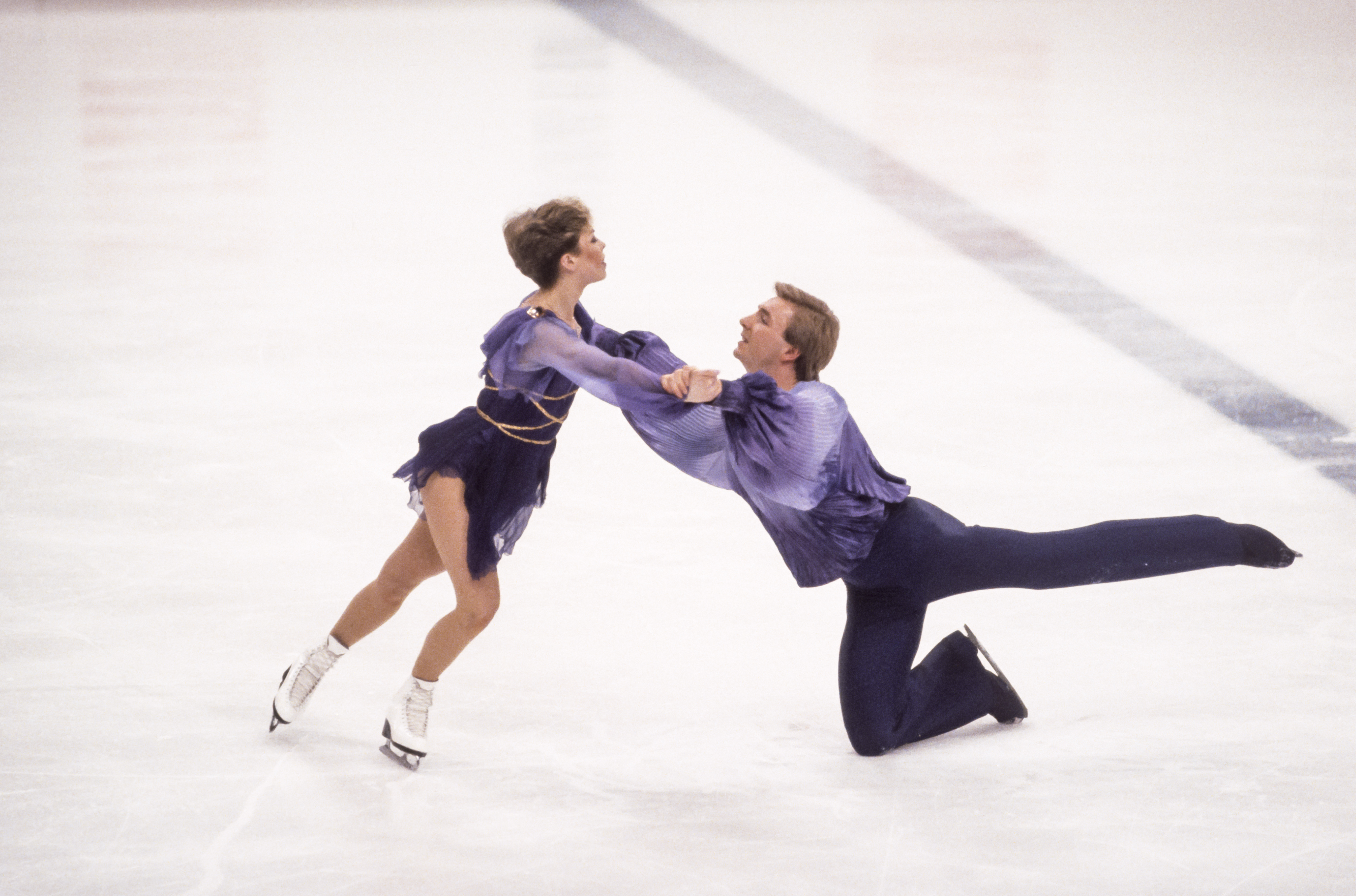 Torvill and Dean perform a dramatic sequence in their iconic Bolero in Sarajevo, 1984. Dean stretches into a deep kneeling line as Torvill drives forward, their connected hold amplifying the routine’s tension and sweep.