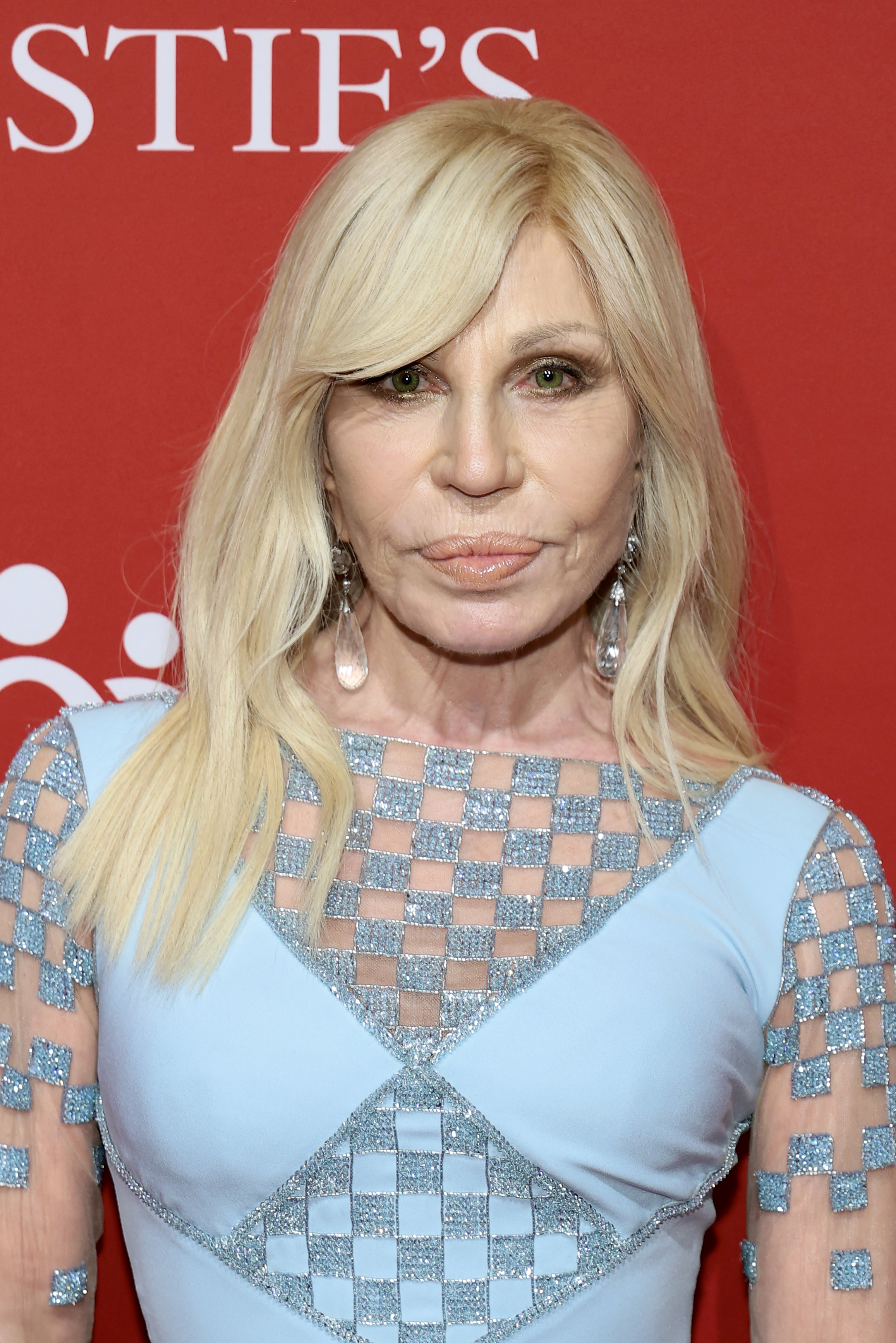 A close-up portrait of Donatella highlights her striking green eyes framed by golden eyeshadow, her platinum blonde hair swept to one side, and large crystal teardrop earrings that brush her shoulders above the gown's embellished neckline.