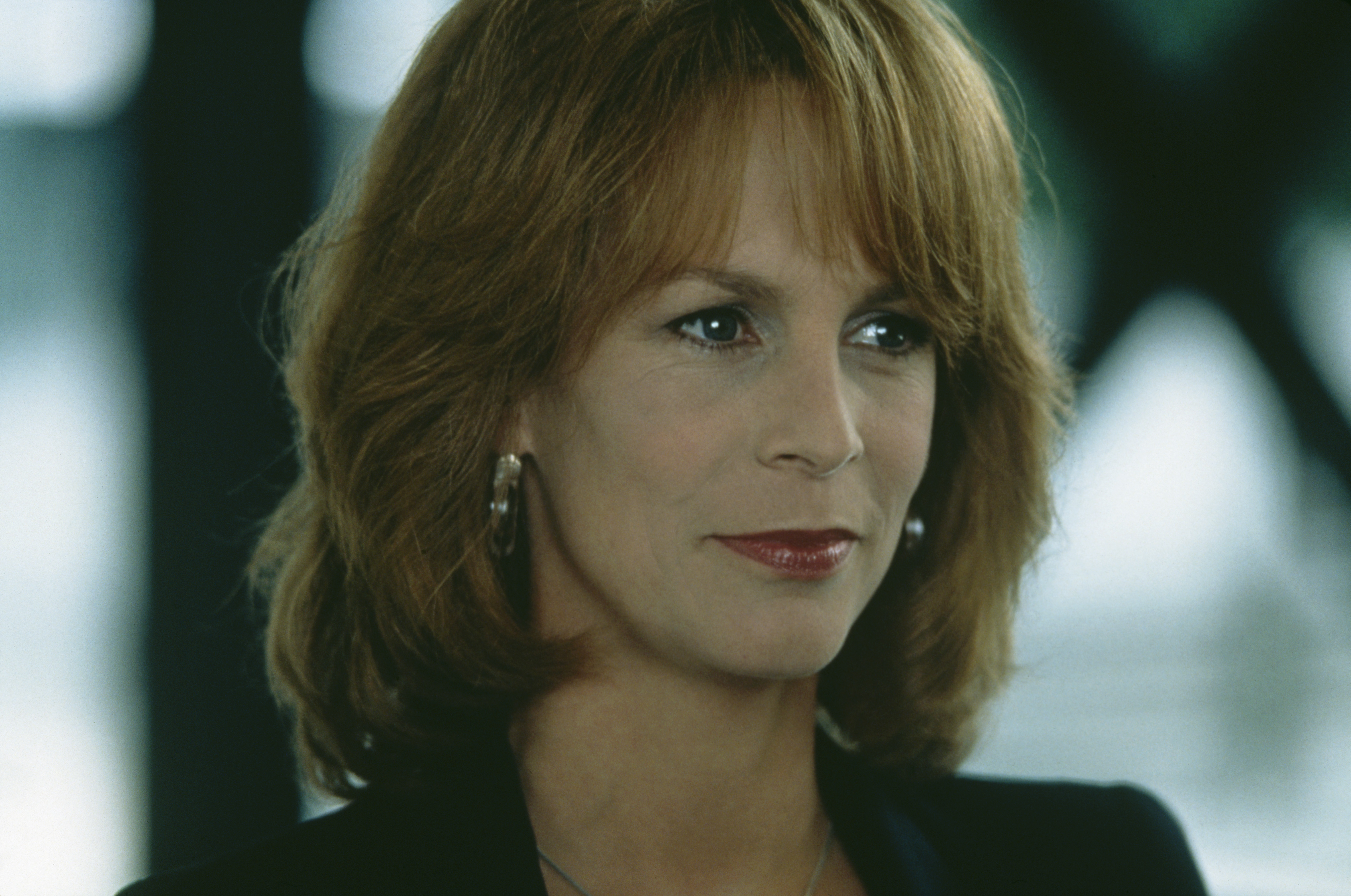 Jamie Lee Curtis appears in character for the 1997 comedy “Fierce Creatures,” wearing voluminous auburn hair with bangs, hoop earrings, and a sharp blazer that matched her poised expression.