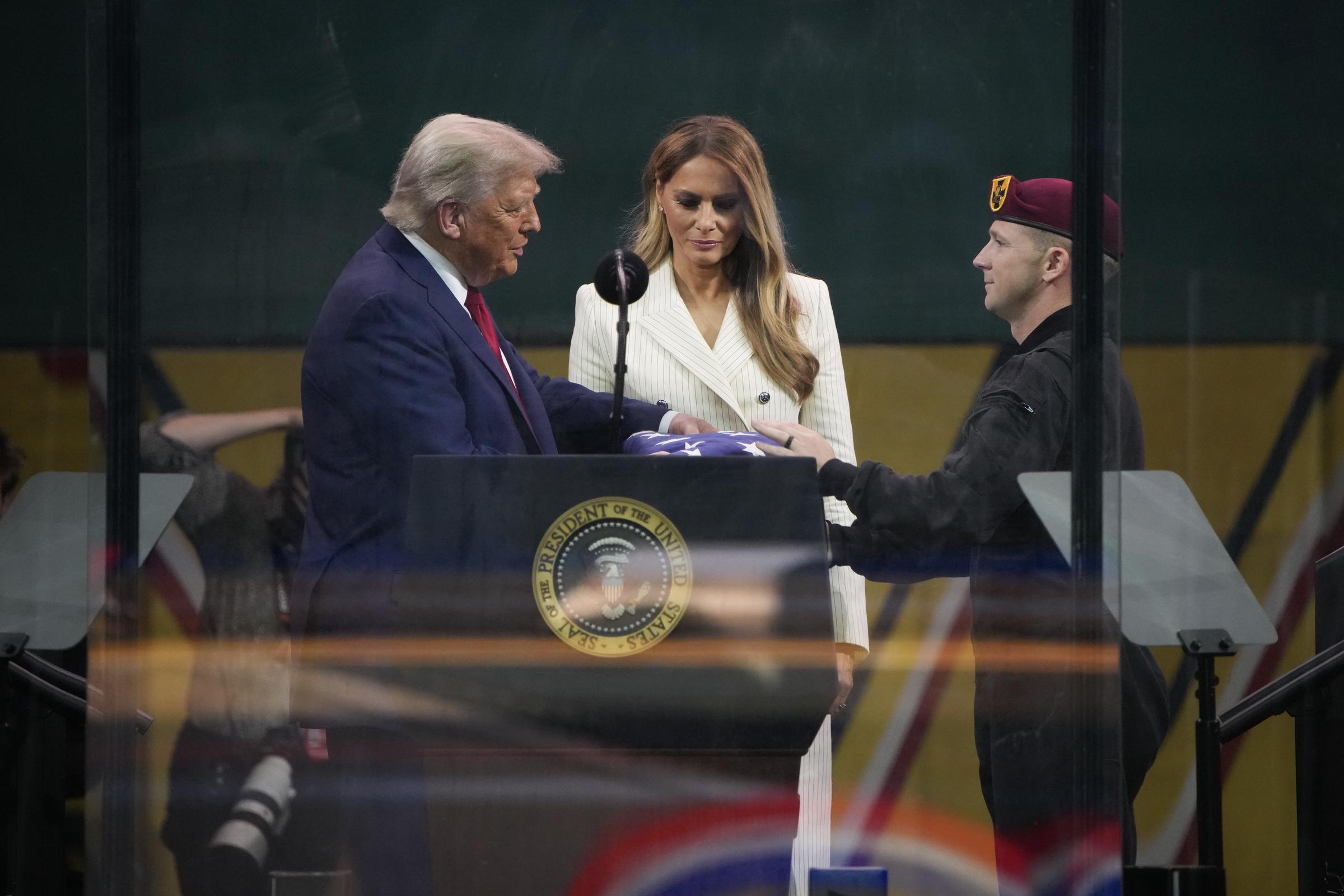 Donald Trump receives a folded American flag from a member of the US Army parachute team as Melania stands beside him. The exchange takes place behind glass at the close of the military ceremony, marking a formal tribute.