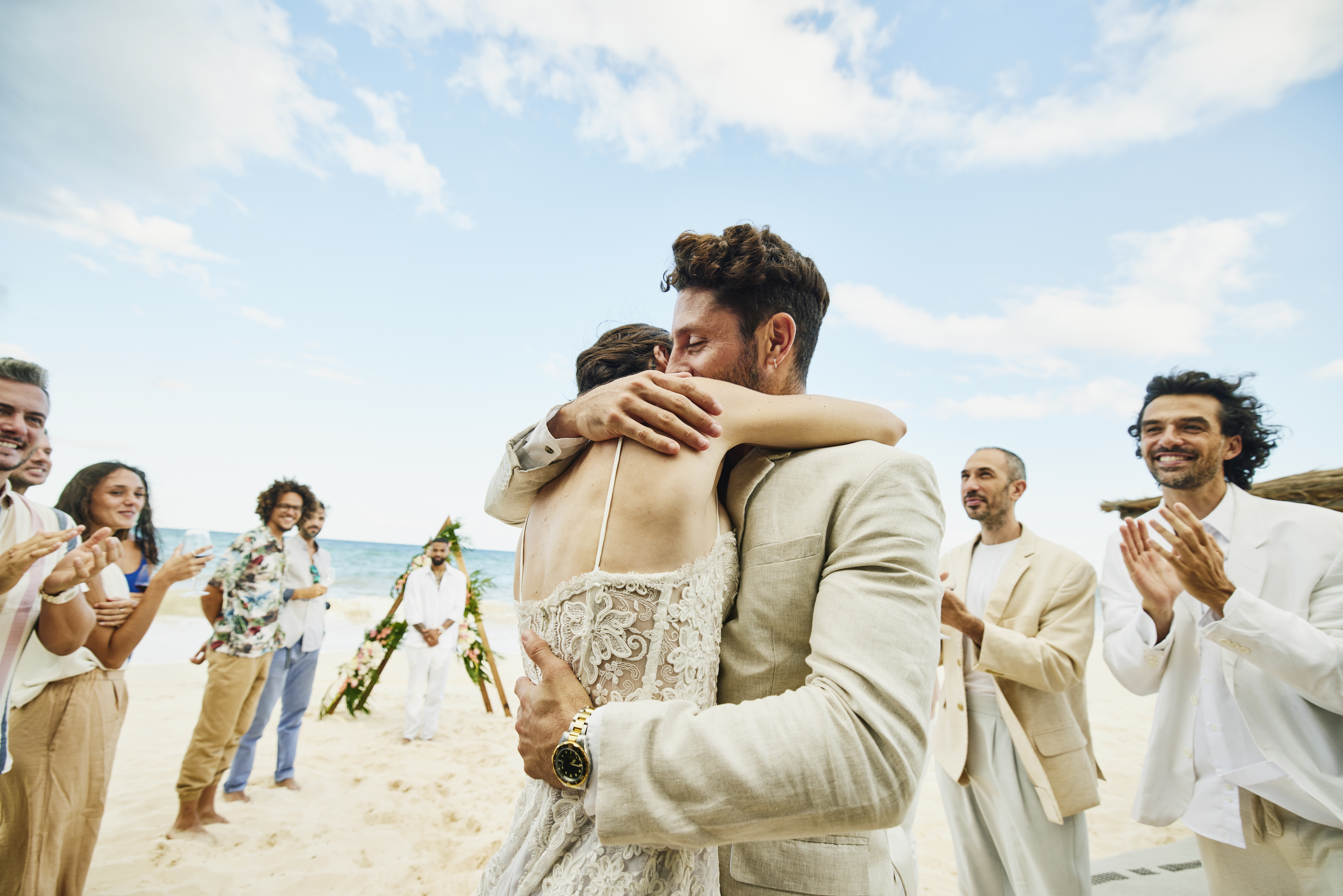 Medium wide shot of bride and groom embracing in front of friends and family celebrating after wedding ceremony on tropical beach | Source: Getty Images