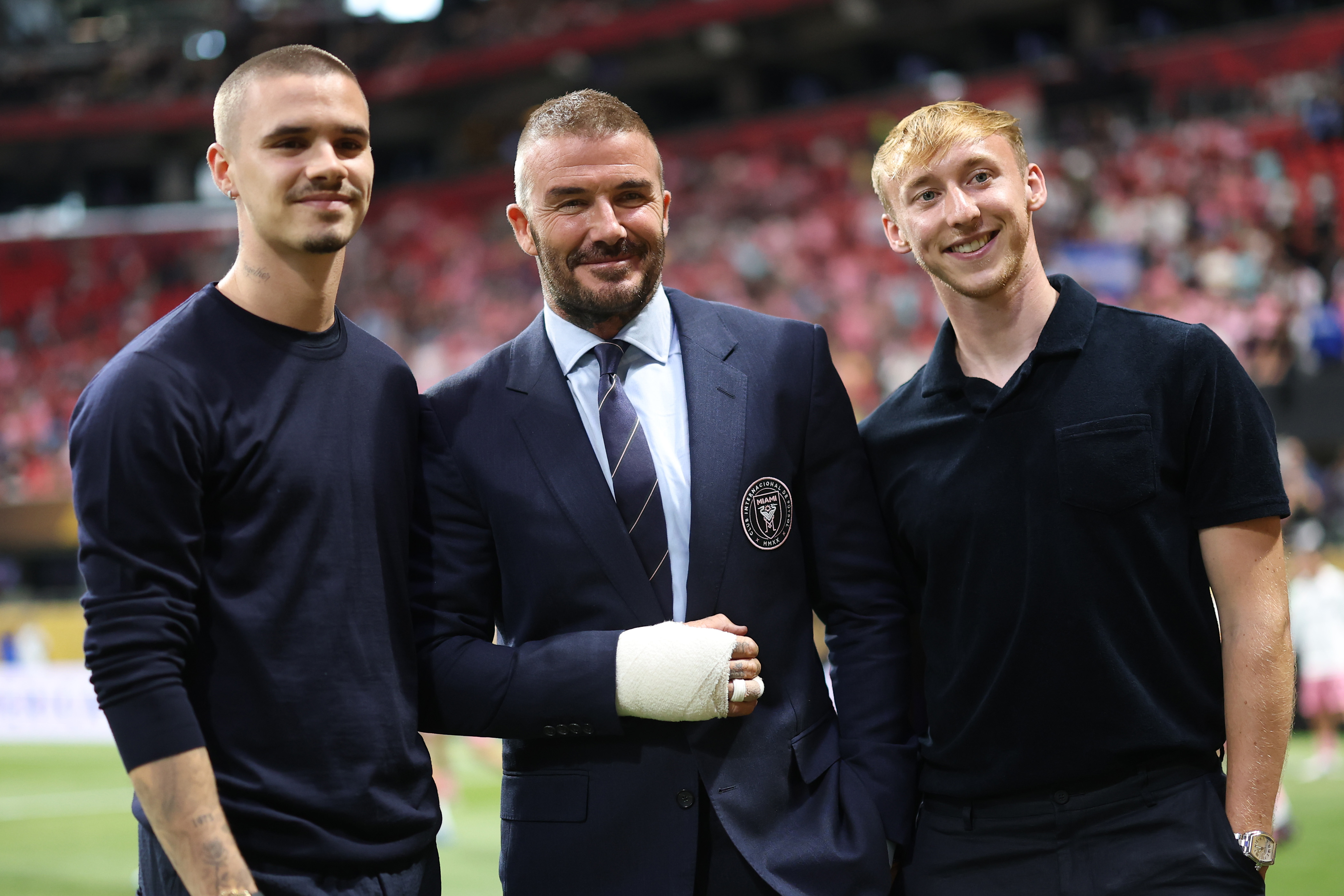 David Beckham poses on the pitch with his son, Romeo Beckham, and Brentford FC’s Max Dickow. His bandaged right hand rests neatly on his suit, but he stays composed and camera-ready before the match.