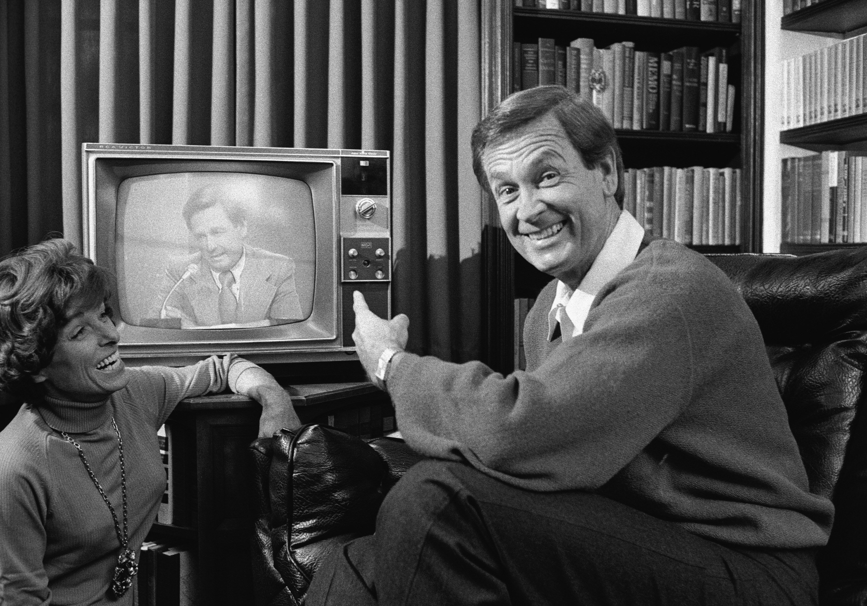 American game show host Bob Barker looks at the camera and smiles while he points to himself on a nearby television screen as his wife Dorothy Jo Barker (1924 - 1981) looks on and laughs, November 4, 1977. | Source: Getty Images