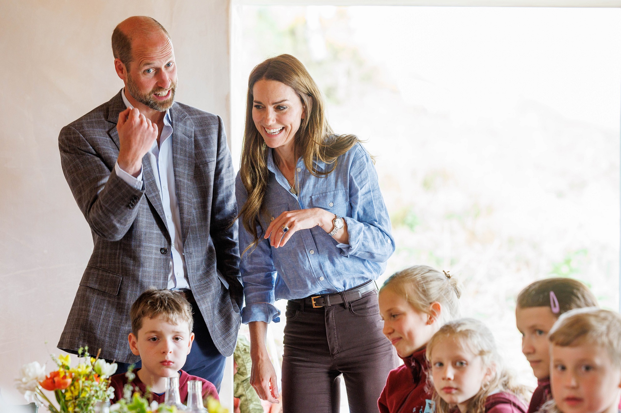 William and Catherine speak to a group of children during a visit to a local croft on the west coast of Mull. Both are smiling and animated, engaging warmly with the young crowd.