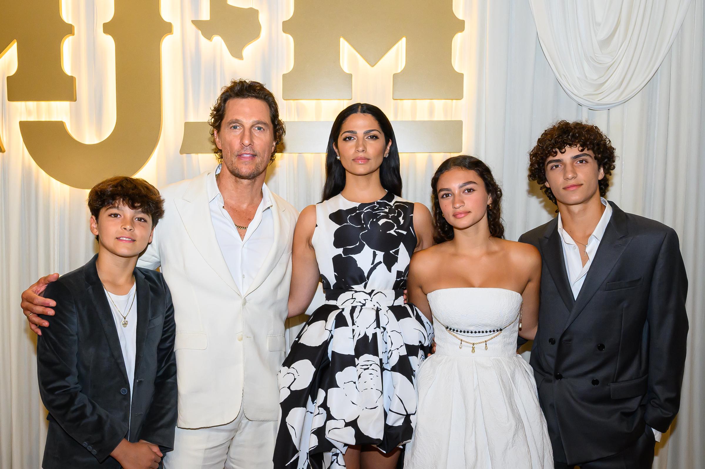 Levi, Livingston, and Vida McConaughey stand closely together in formal attire at another MJM Gala event. Levi wears a suit, Livingston a blazer, and Vida a fitted dress. Their parents, Matthew McConaughey and Camila Alves McConaughey stand behind them facing forward on the event backdrop.