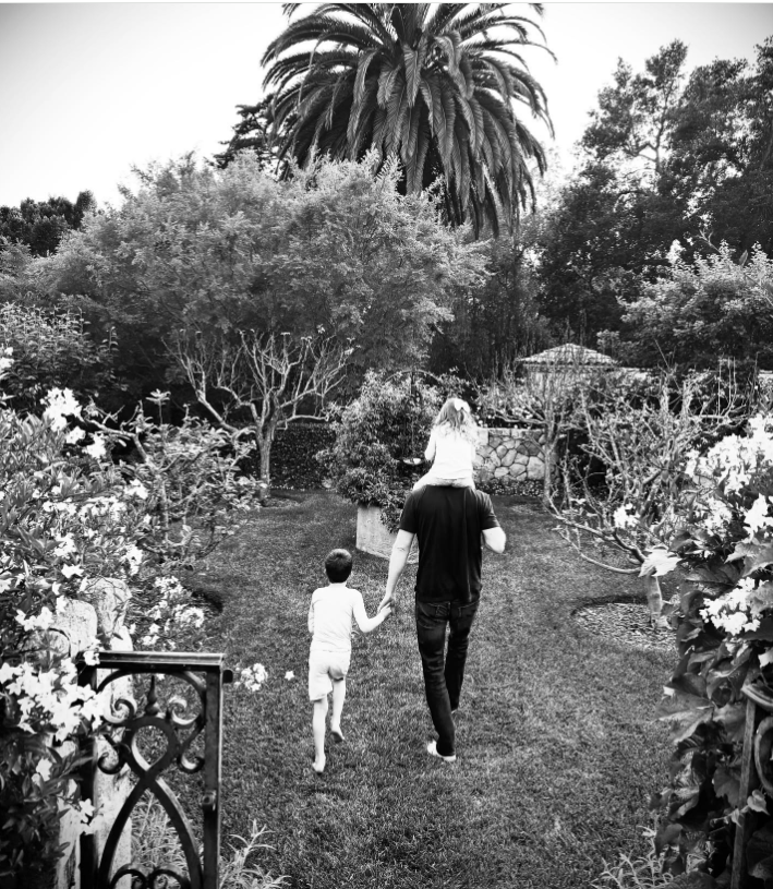 Prince Harry strolls through a manicured garden with his children, carrying Princess Lilibet on his shoulders while holding Prince Archie's hand. | Source: Instagram/meghan