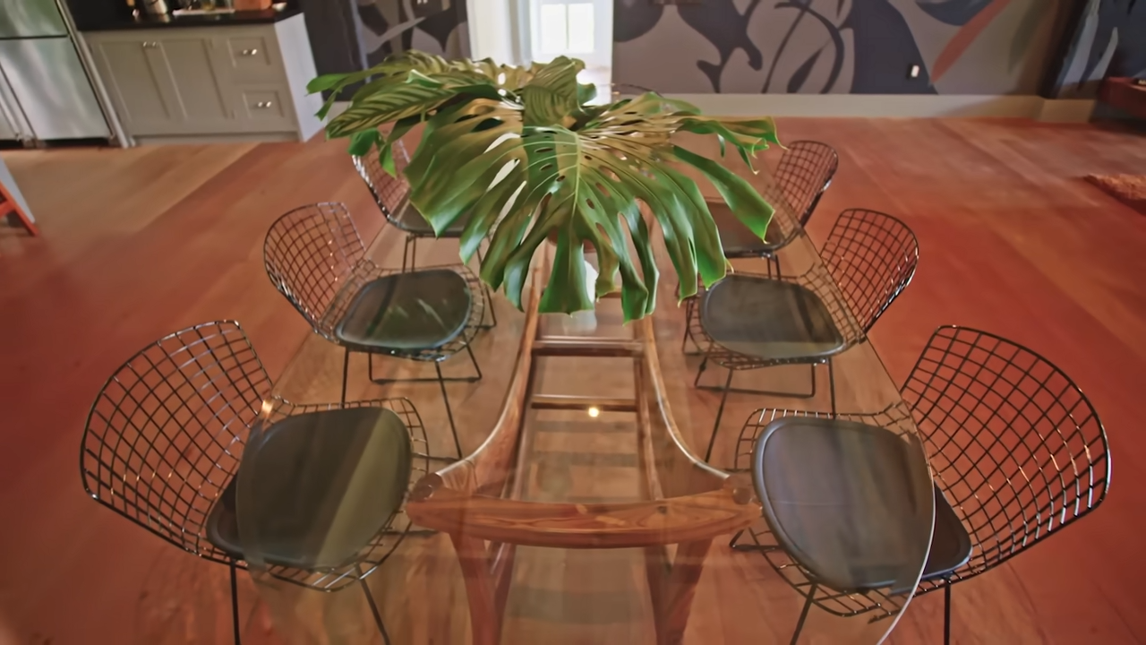A sleek, glass-topped dining table with a sculptural wood frame is paired with minimal wire chairs and topped with a giant monstera leaf centerpiece. | Source: YouTube/@Archdigest