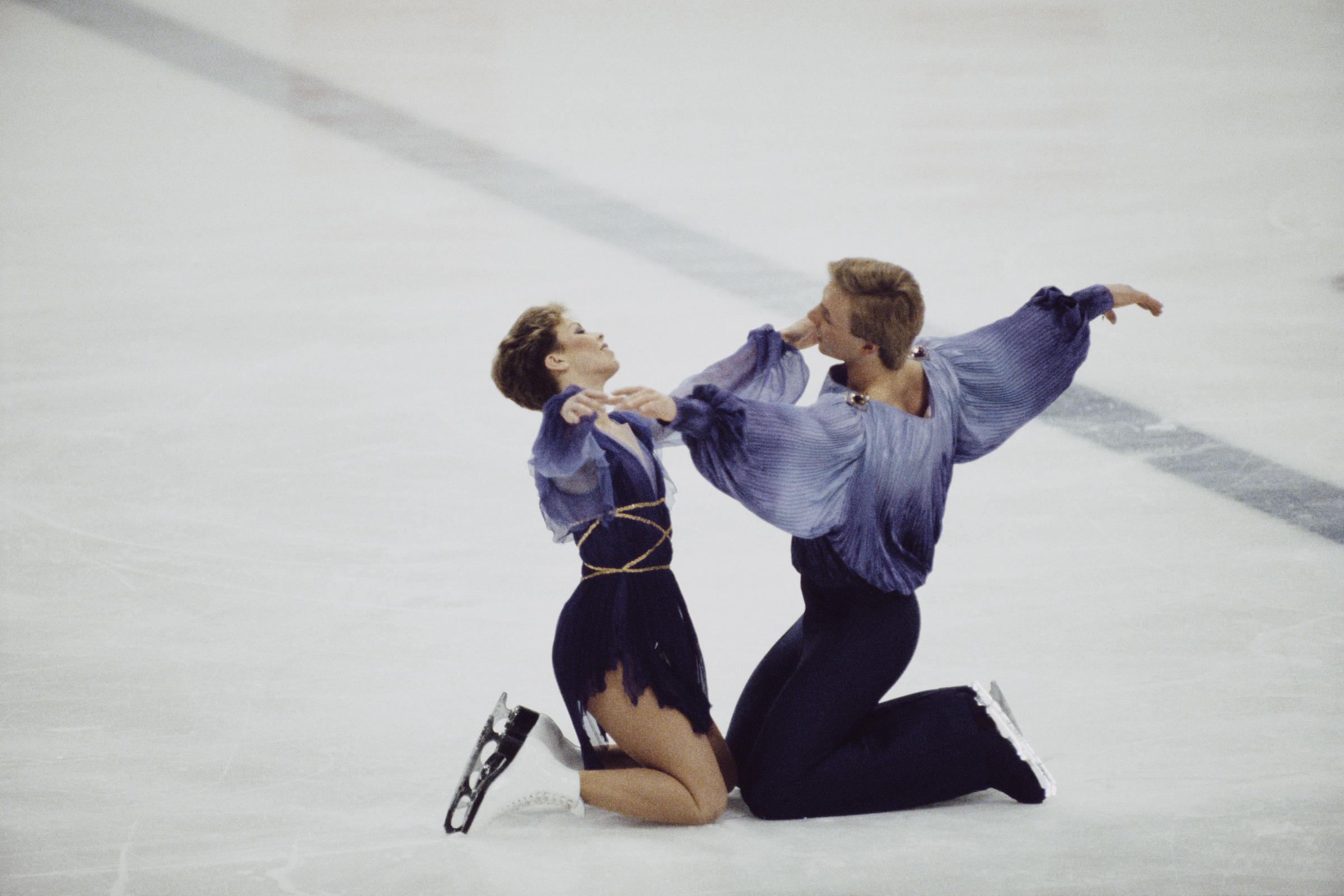 Jayne Torvill and Christopher Dean kneel facing one another during their gold medal-winning Bolero at the 1984 Winter Olympics. Arms extended in mirrored lines, they hold a moment of poised stillness that heightens the routine’s dramatic intensity.