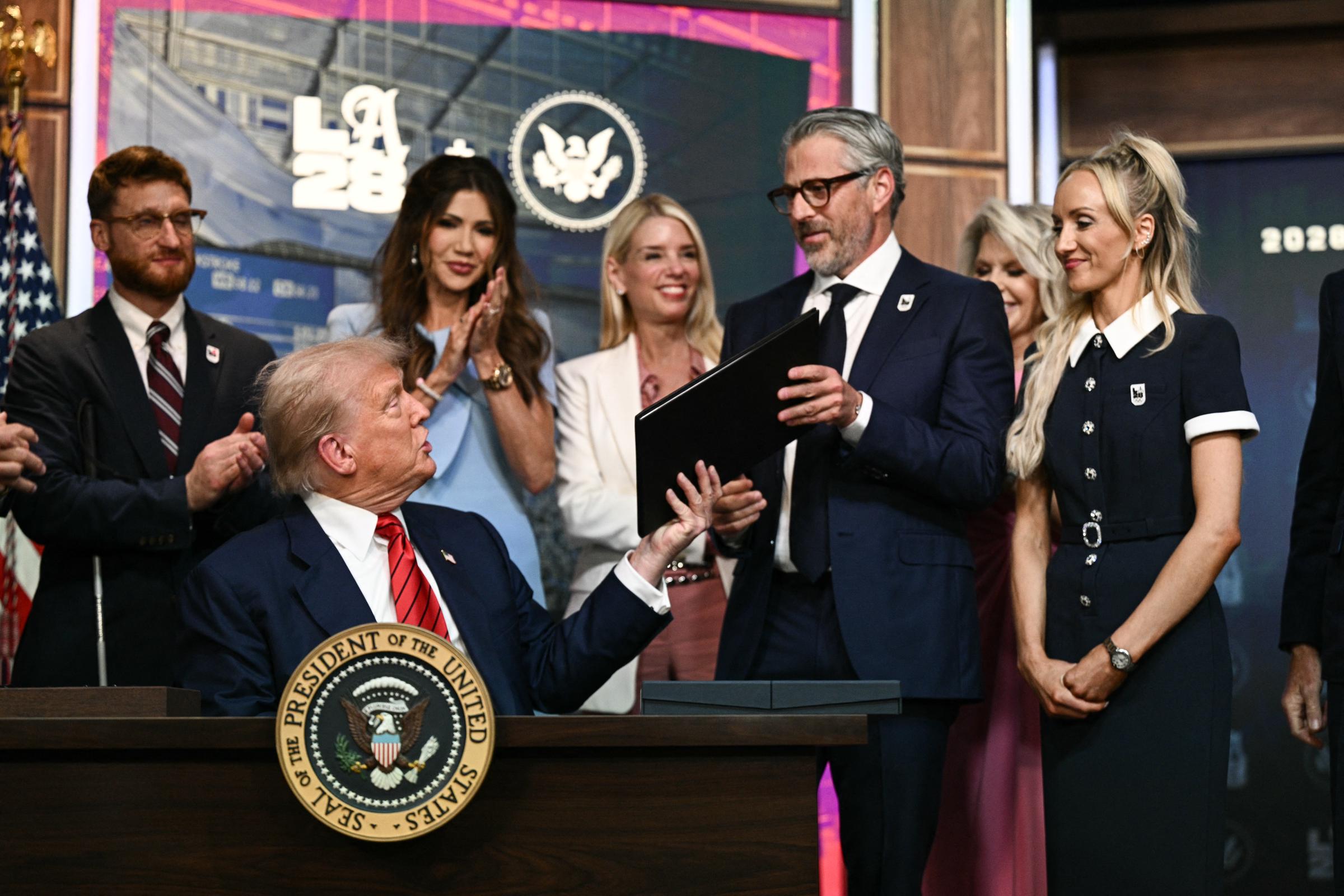 Donald Trump appears to speak as he hands LA 2028 Chairman Casey Wasserman a copy of the signed executive order, while attendees look on with smiles and applause.