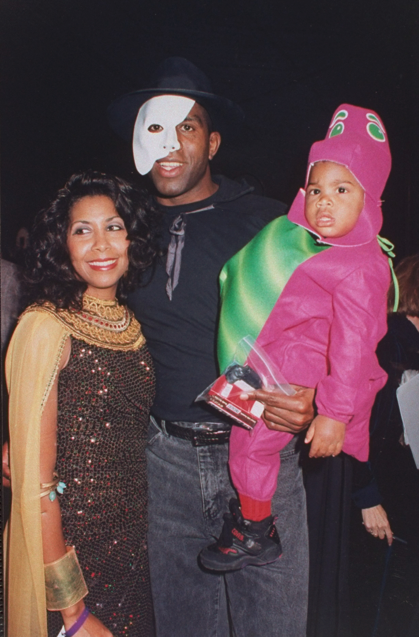 Magic Johnson, wearing a "Phantom of the Opera" mask, holds his son Earvin III dressed as Barney alongside his wife Cookie in an Egyptian costume at the Halloween on the Green AIDS fundraiser at Tavern on the Green on October 31, 1993, in New York City