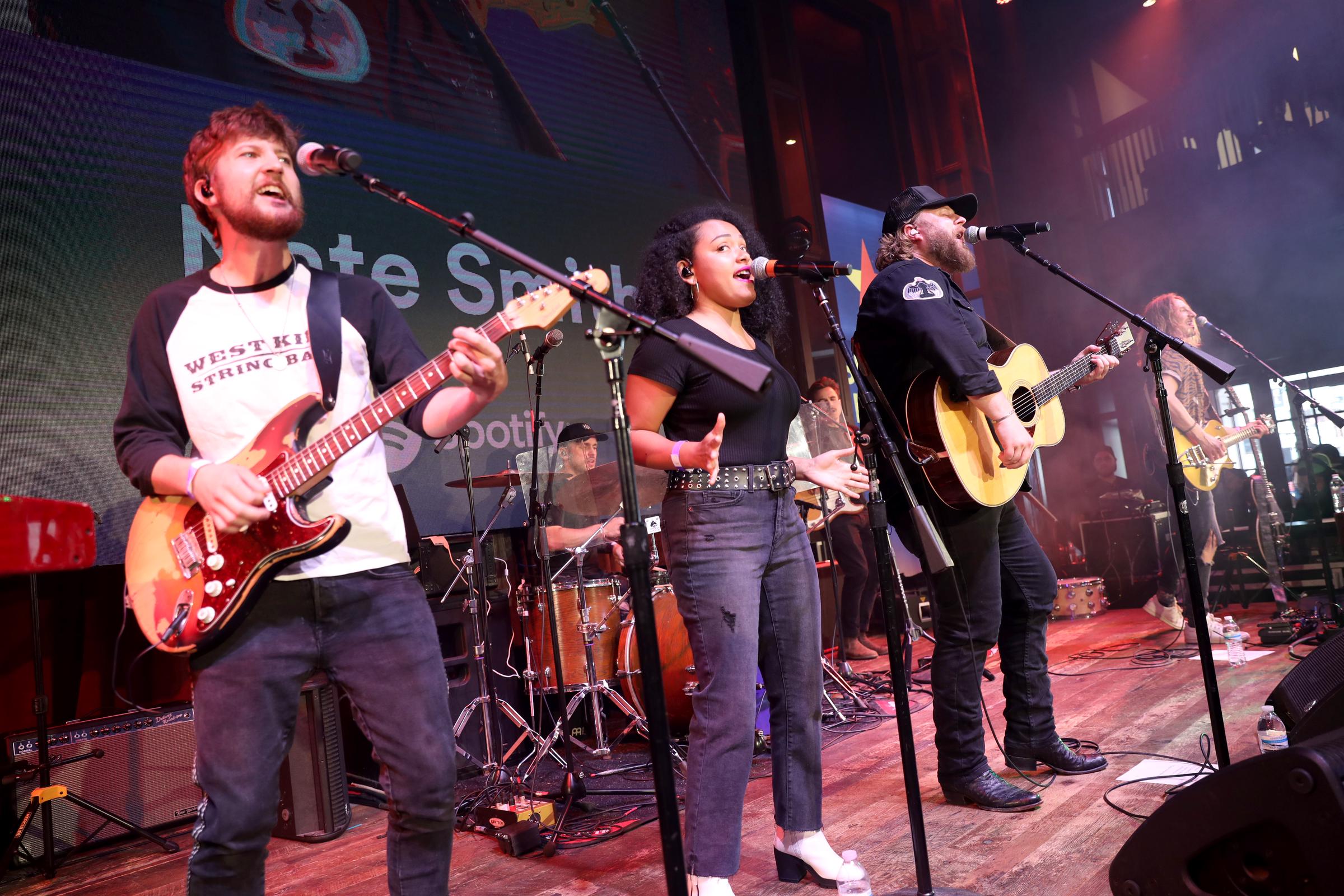 Nate Smith performs alongside his band, singing and playing acoustic guitar as the crowded stage comes alive with musicians, backing vocals, and colorful lights.