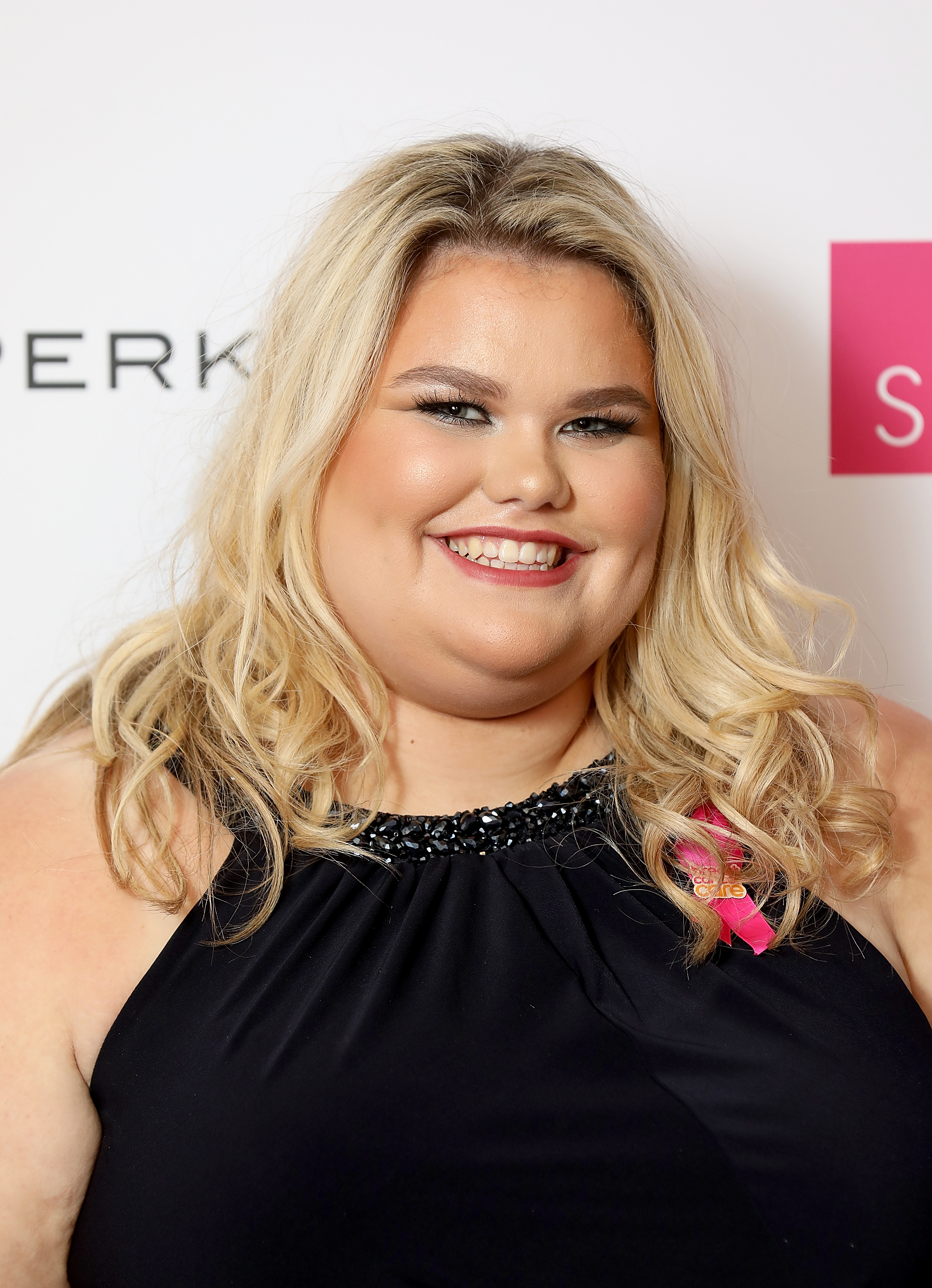 A confident Tapper, her blonde waves styled loosely around her face, flashes a wide smile at the Breast Cancer Care London Fashion Show in October 2018. She wears a black embellished halterneck dress and a pink charity ribbon pinned to her chest.