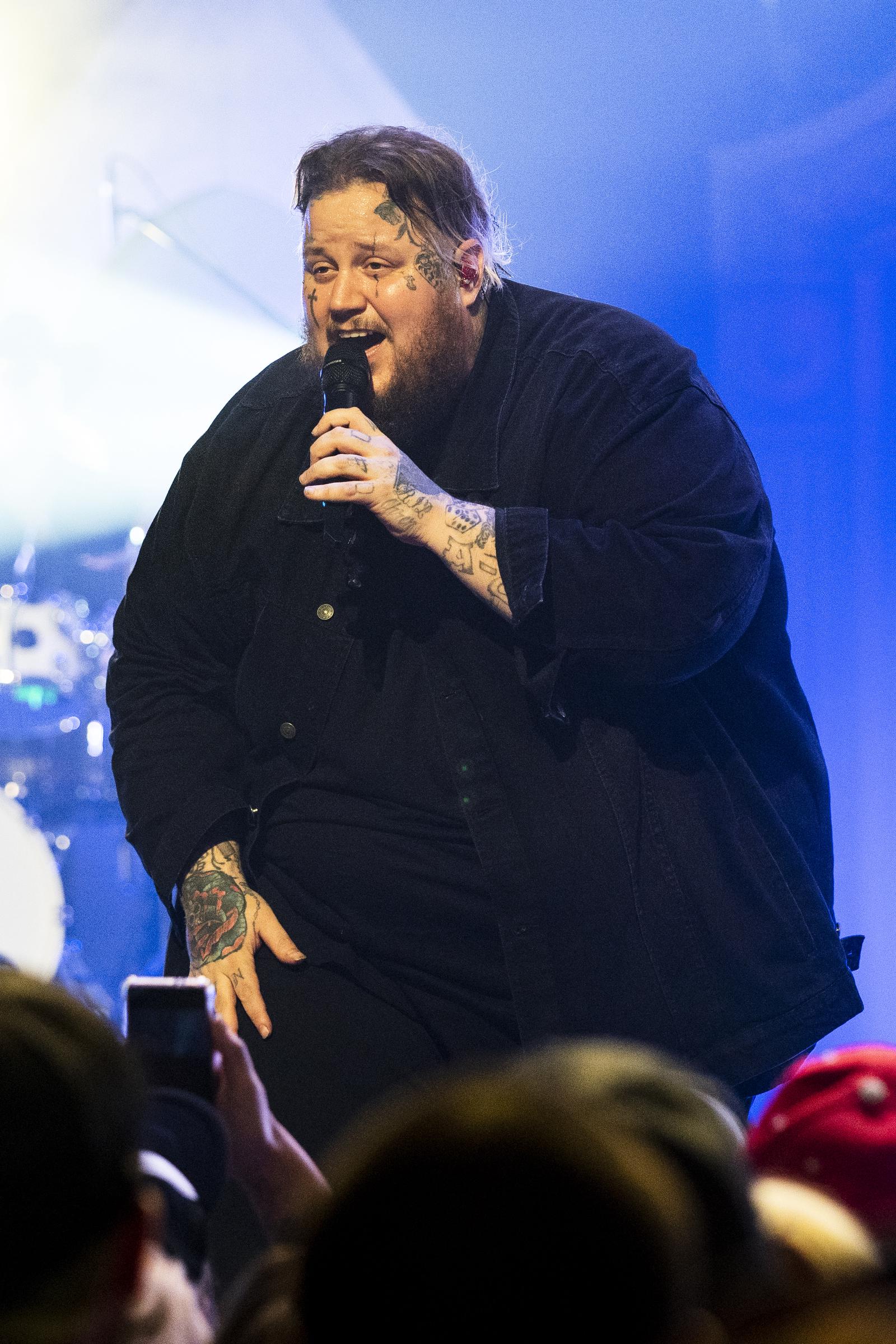 Jason "Jelly Roll" DeFord captured mid-performance, leaning forward with a warm, engaging smile as he sings into the microphone at the Orpheum Theater in New Orleans, Louisiana. Dressed in an all-black ensemble, his tattoos are prominently visible on his hands and face, illuminated by the stage lights. The vibrant atmosphere and his expressive energy reflect a strong connection with the audience.