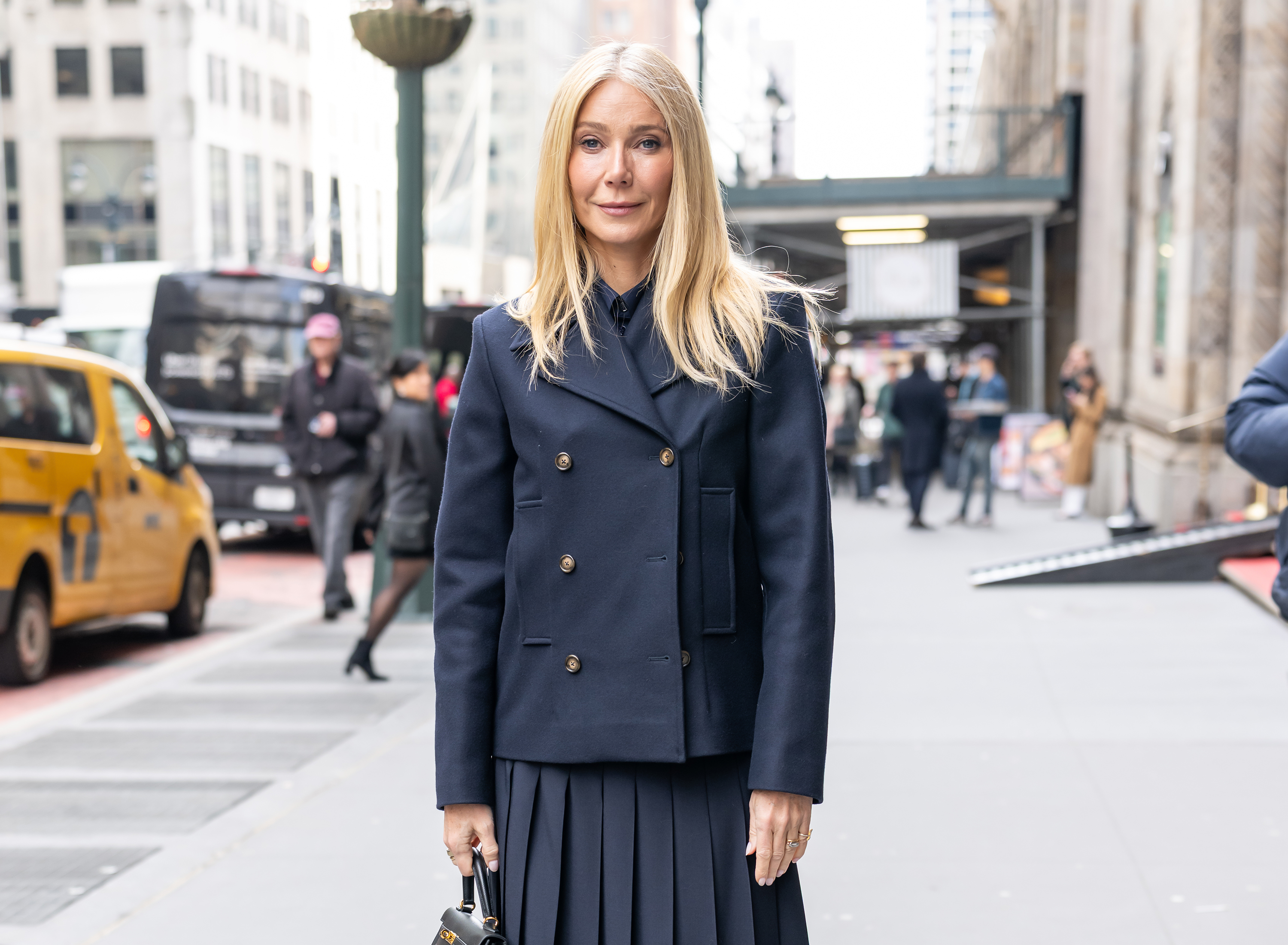Gwyneth Paltrow faces forward on a busy New York street, holding a black Hermès Kelly bag, her pleated skirt falling neatly as her calm expression and minimal styling stand out.