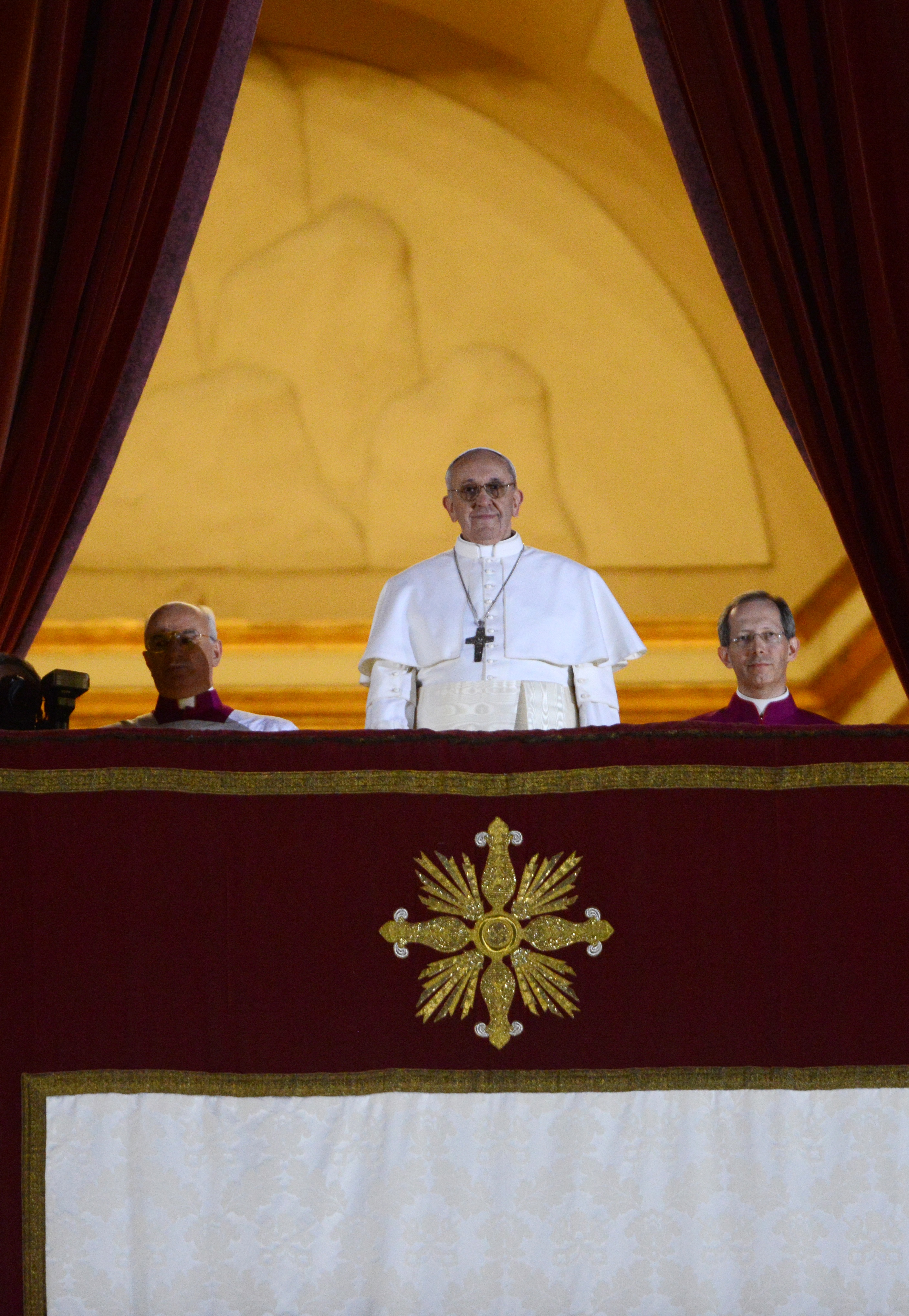 Pope Francis stands beneath deep red drapes, framed by golden light and solemnly gazing out in white papal robes and a silver cross
