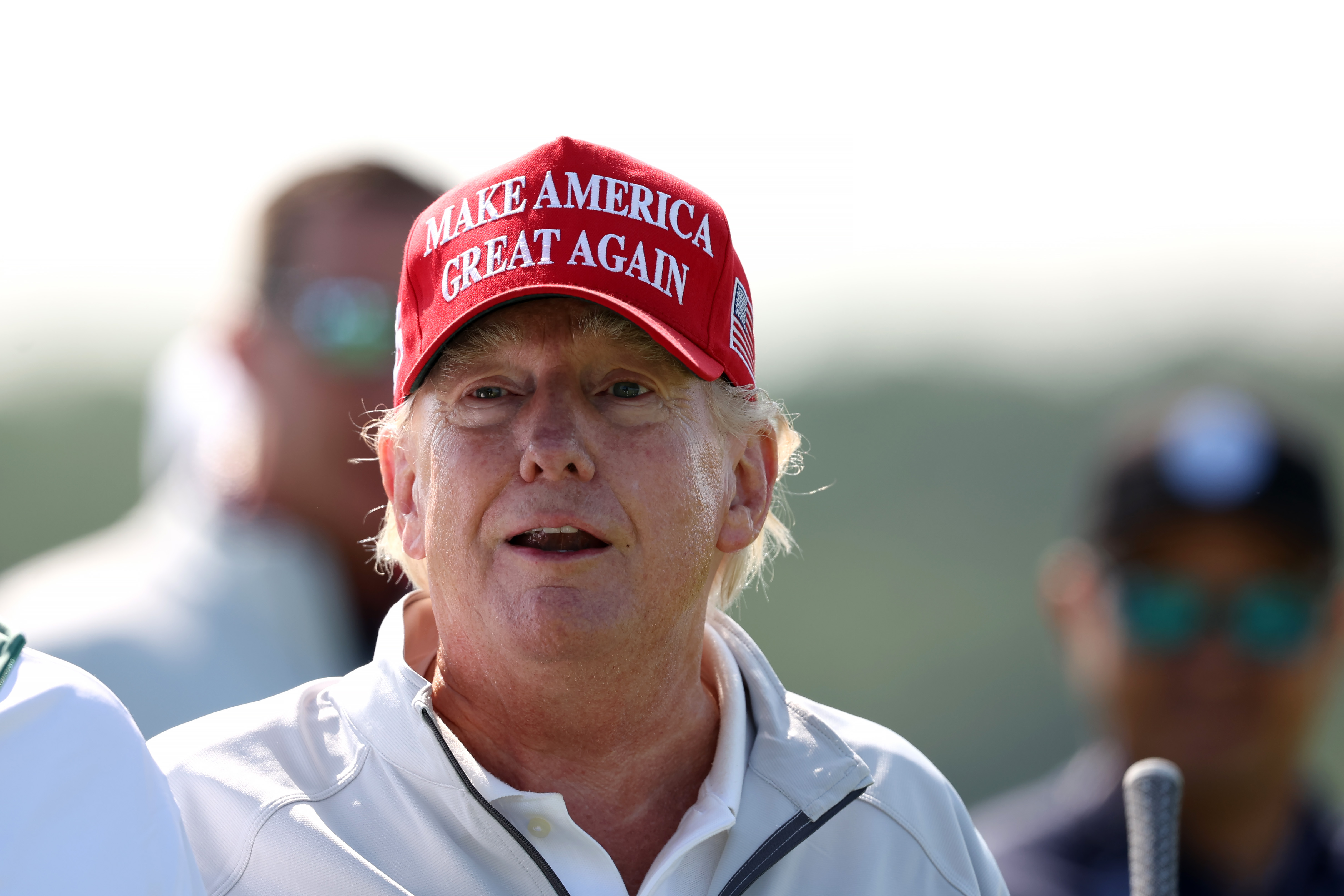 At the LIV Golf Invitational in Sterling, Virginia, on May 25, Donald Trump stands in sunlight wearing a red "Make America Great Again" cap and a white collared jacket. His cheeks and nose appear deeply flushed, while his neck looks much lighter.