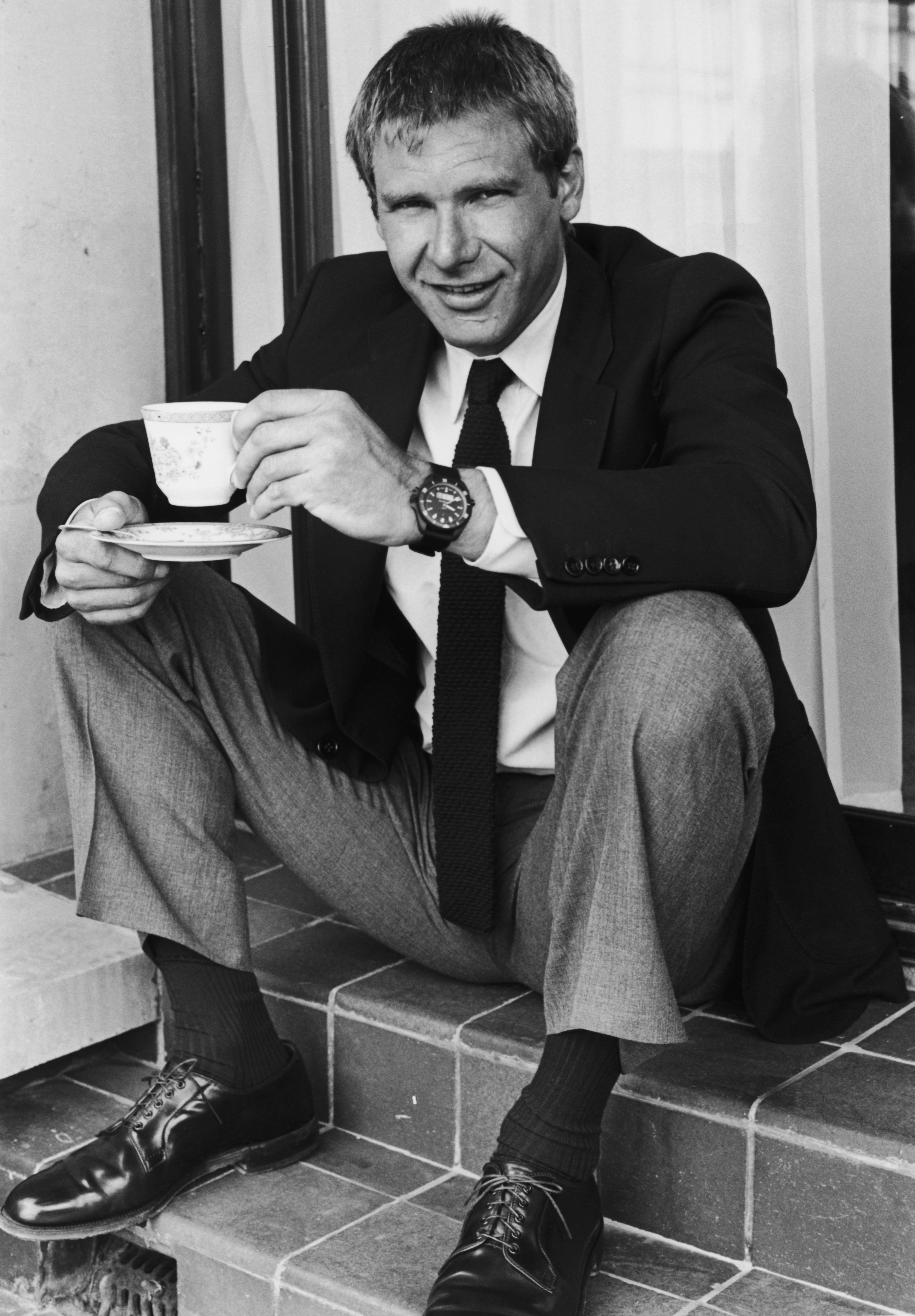 Harrison Ford sitting on steps, smiling while holding a teacup and saucer in circa 1984. | Source: Hulton Archive/Getty Images
