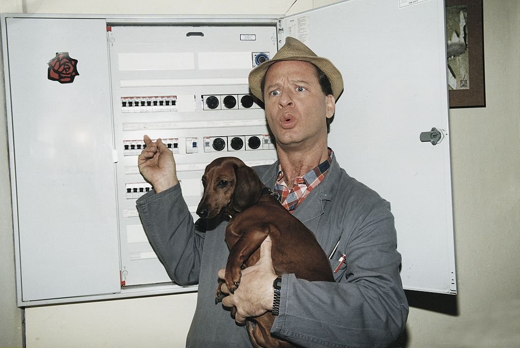 Tom Gerhardt vor einem Sicherungskasten als "Hausmeister Krause" mit Hut und dem Dackel 'Bodo' auf dem Arm. (Foto von: Moenkebild) I Quelle: Getty Images