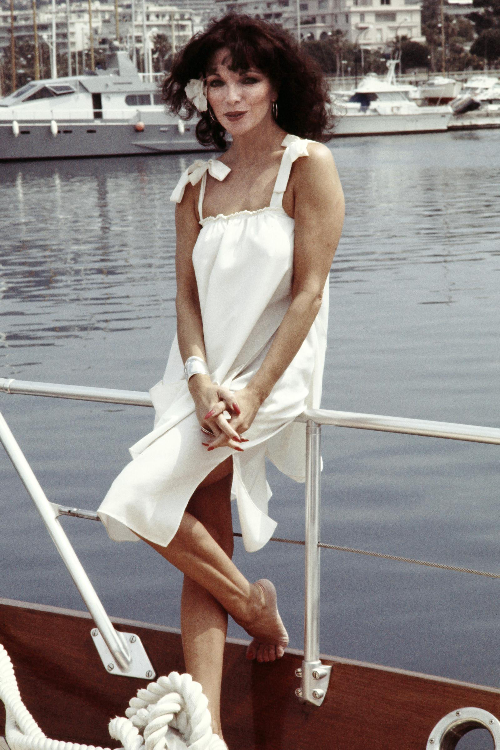 Joan Collins poses barefoot on the deck of a boat during the Cannes Film Festival. She wears a flowing white sundress with tied shoulder straps and a large flower pinned in her curled hair. Her nails are painted red, and she smiles softly with the marina and yachts behind her.