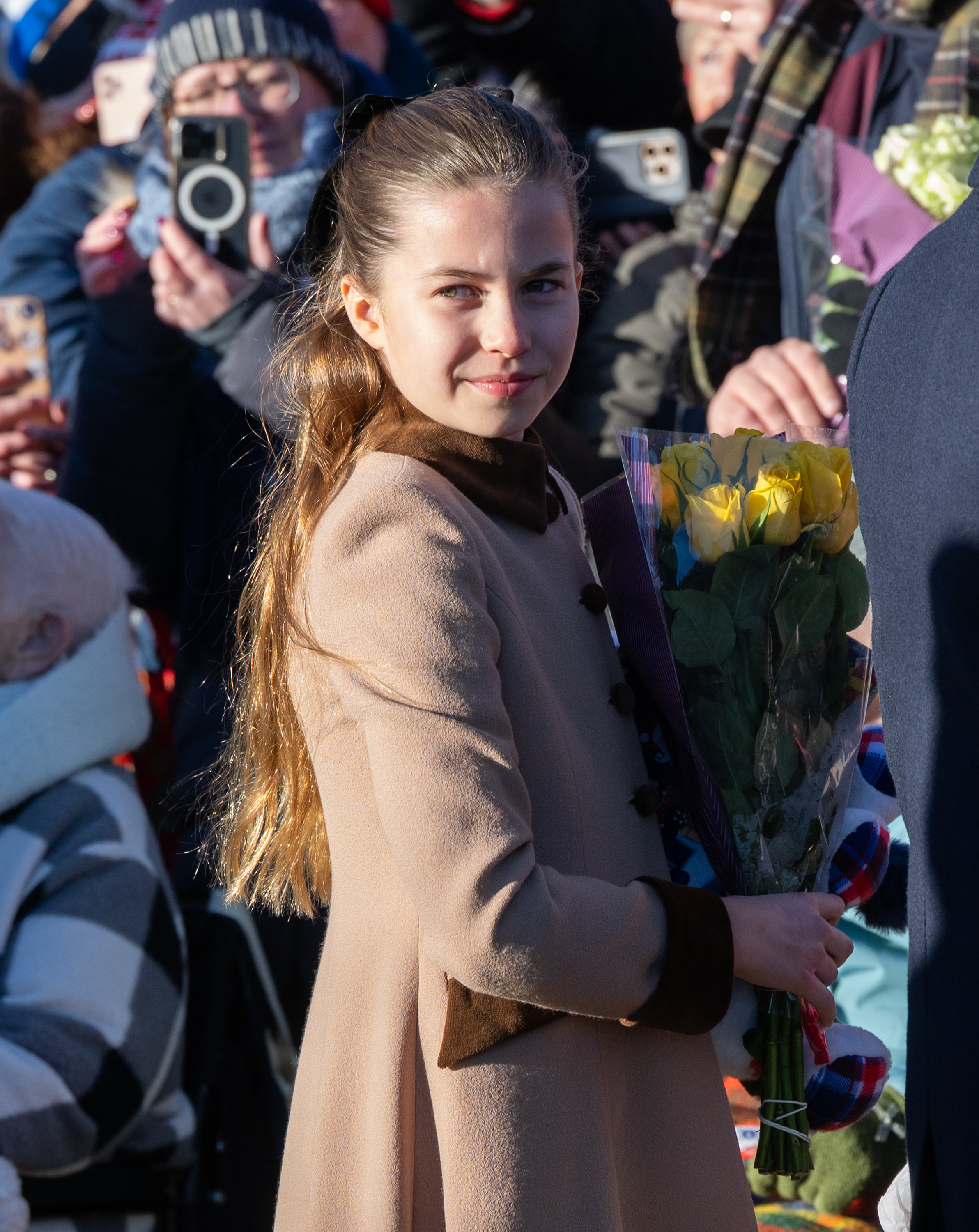 Charlotte glances back over her shoulder with a quiet smile, clutching a bouquet of yellow roses gifted by a wellwisher, as she works the crowd at the Sandringham Christmas Morning Service in December 2025.