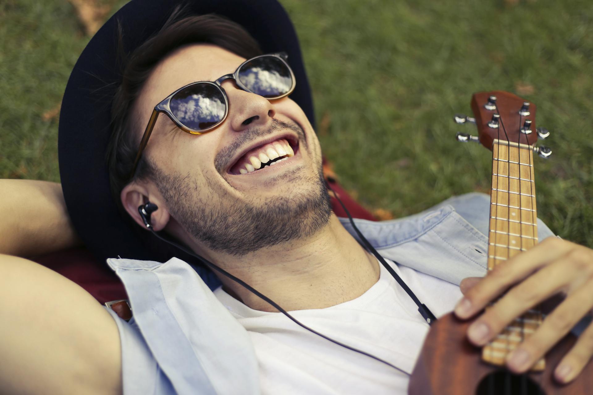 A man wearing a hat and sunglasses lying on a grass field listening to music and holding a guitar | Source: Pexels