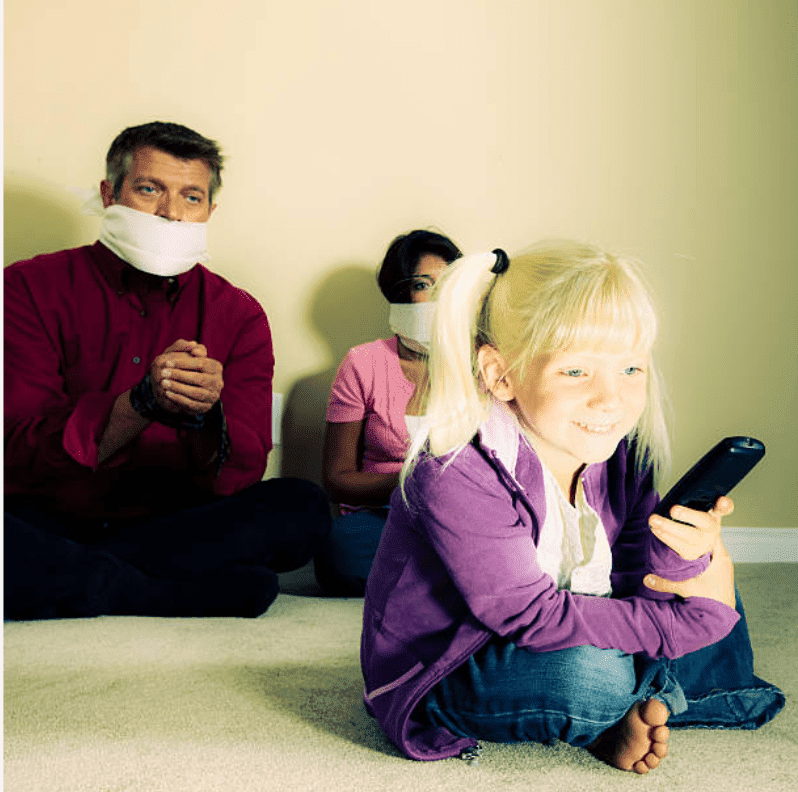 Little blonde girl smiles holding the remote and watching television, while her parents a tied up behind her | Source: Getty Images