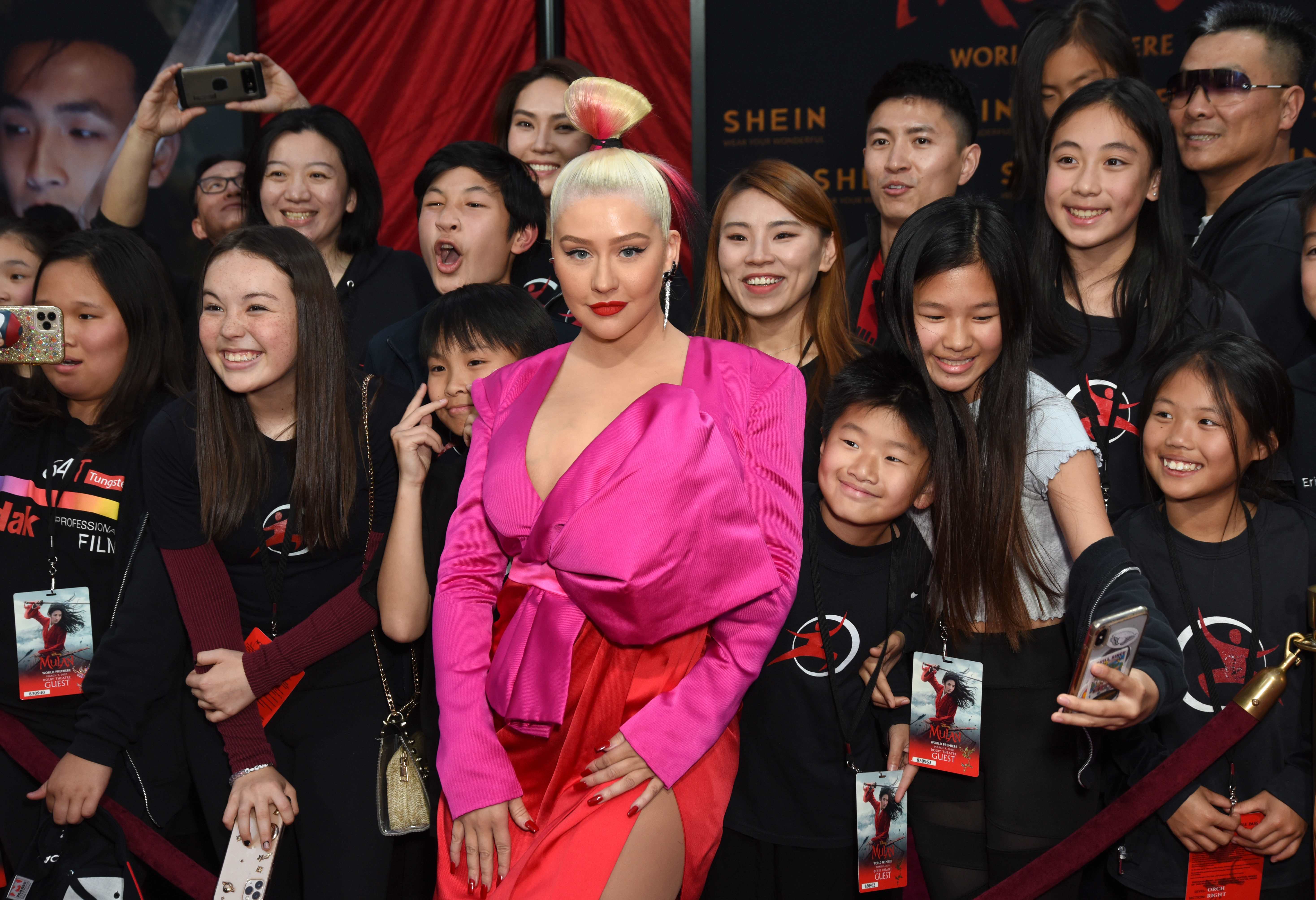 Christina Aguilera poses with fans at the world premiere of Disney's "Mulan" at the Dolby Theatre in Hollywood in March 2020. She wears a bold pink and red gown with a dramatic oversized bow and thigh-high slit, complemented by a slick high bun dipped in pink — a striking look that blended high fashion with playful color.