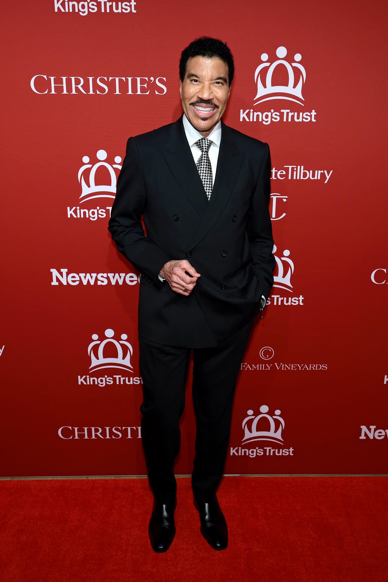 Gala co-chair Lionel Richie beams on the red carpet in a sharp double-breasted black suit over a crisp white shirt, finished with a black-and-white houndstooth tie and polished black dress shoes, his signature warm smile firmly in place.