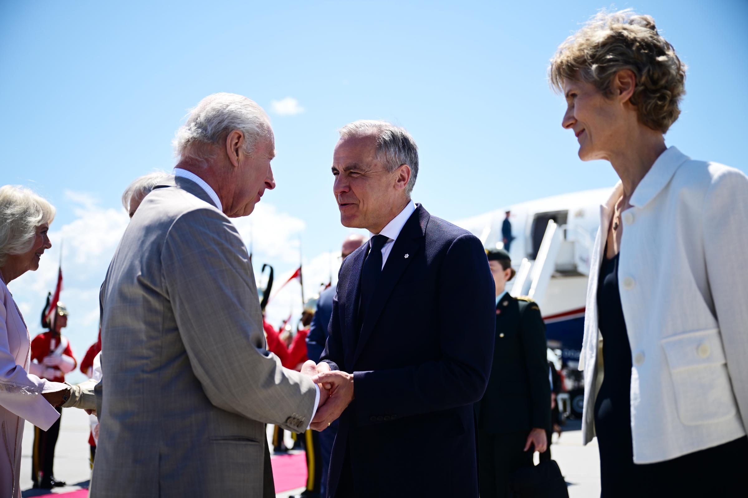 A shot focuses on King Charles III as he is greeted with both hands by Prime Minister Mark Carney, while Diana Fox Carney looks on, with Queen Camilla slightly out of focus in the background.